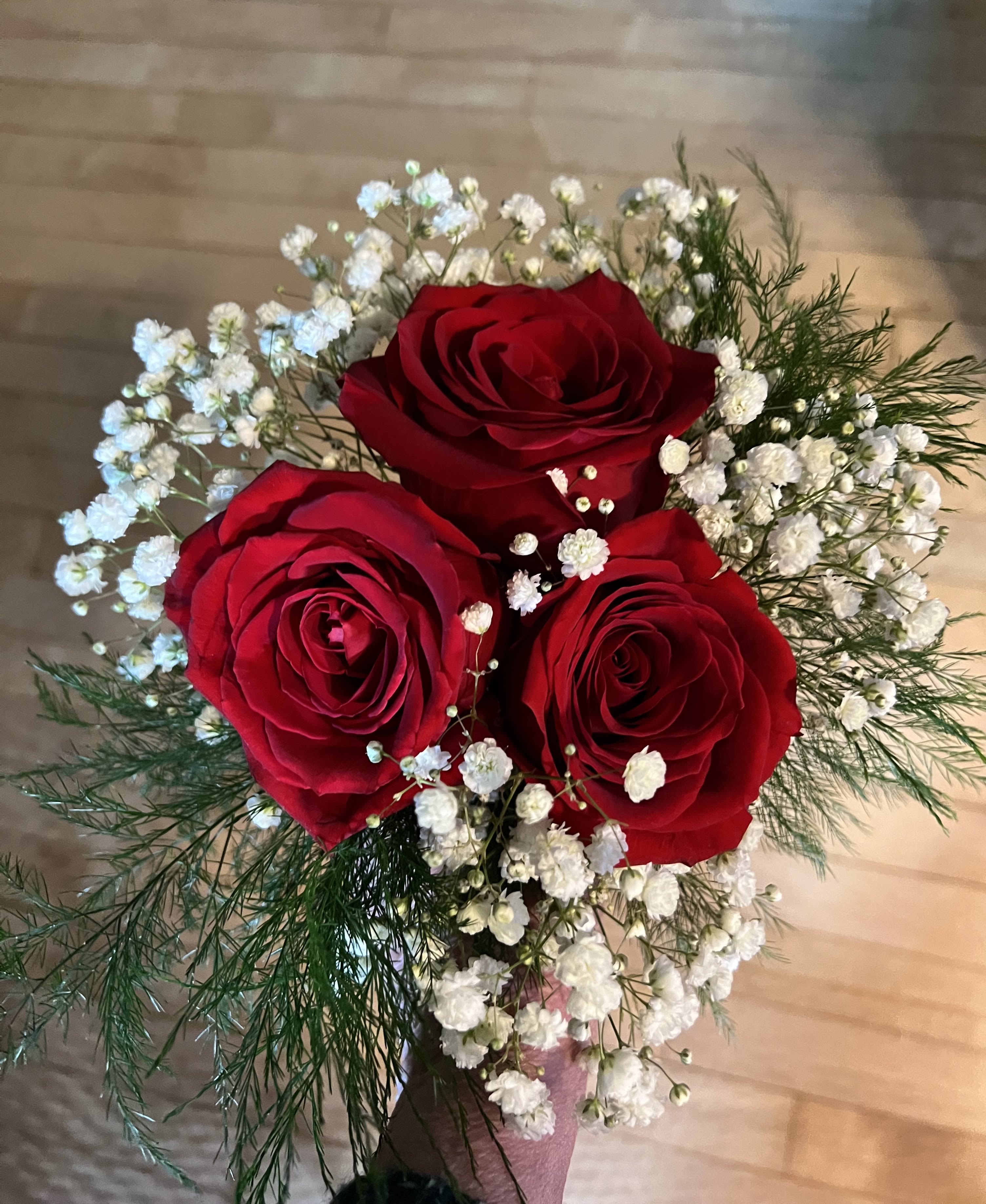 Petite Bouquet - 3 Roses surrounded with filler and greenery. Hand wrapped and tied with a ribbon.  Deluxe Bouquet would contain 6 roses. Premium would contain 9 roses. Your choice of ribbon color and rose color.