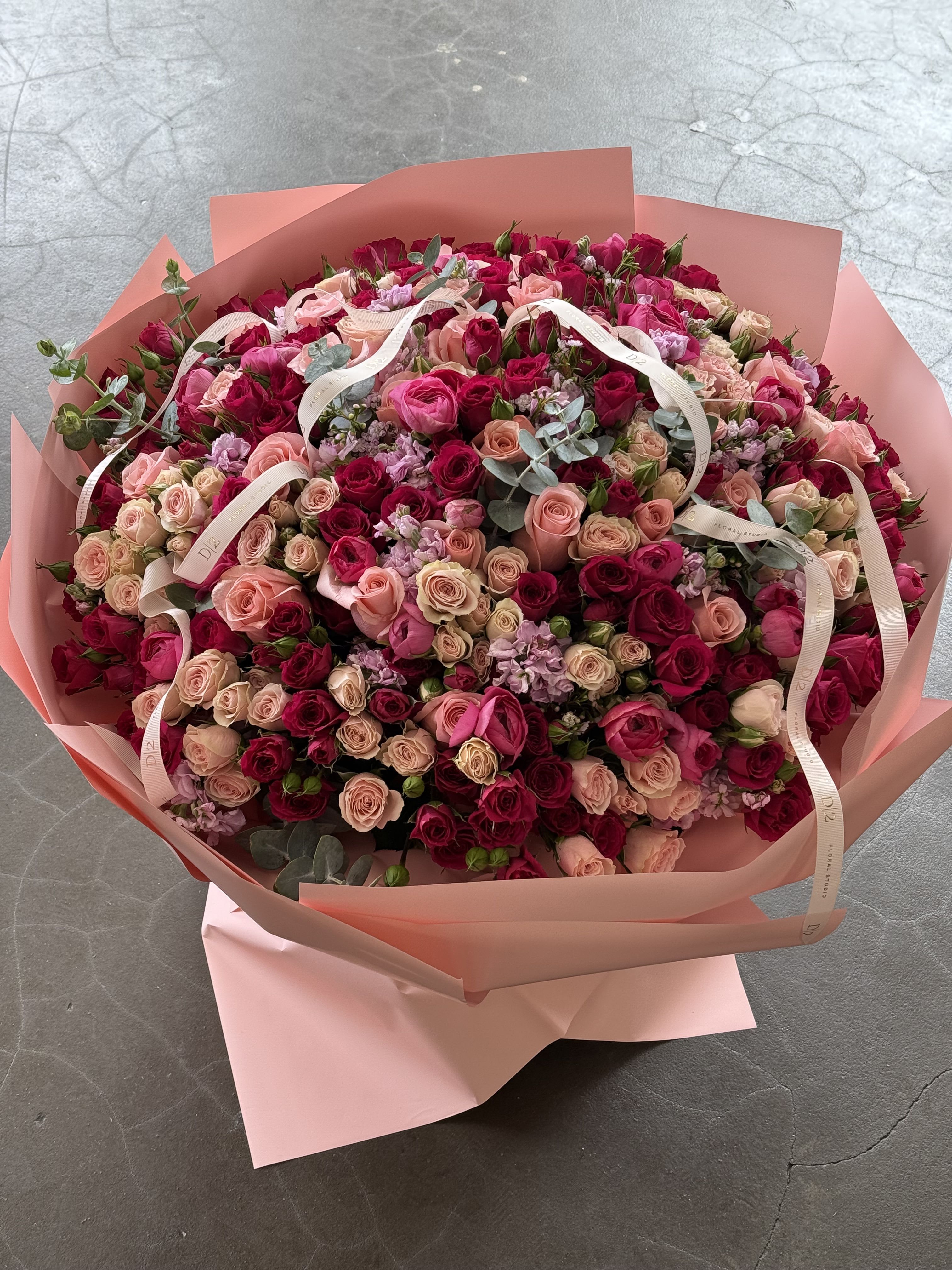 Tereza - This stunning, voluminous bouquet features a rich blend of deep fuchsia, blush, and soft pink roses, accented with delicate lavender blooms and sprigs of eucalyptus for texture and contrast. Wrapped in elegant pink paper and tied with signature ivory “D|2” ribbons, this luxurious arrangement radiates romance and sophistication—perfect for grand gestures and special celebrations.