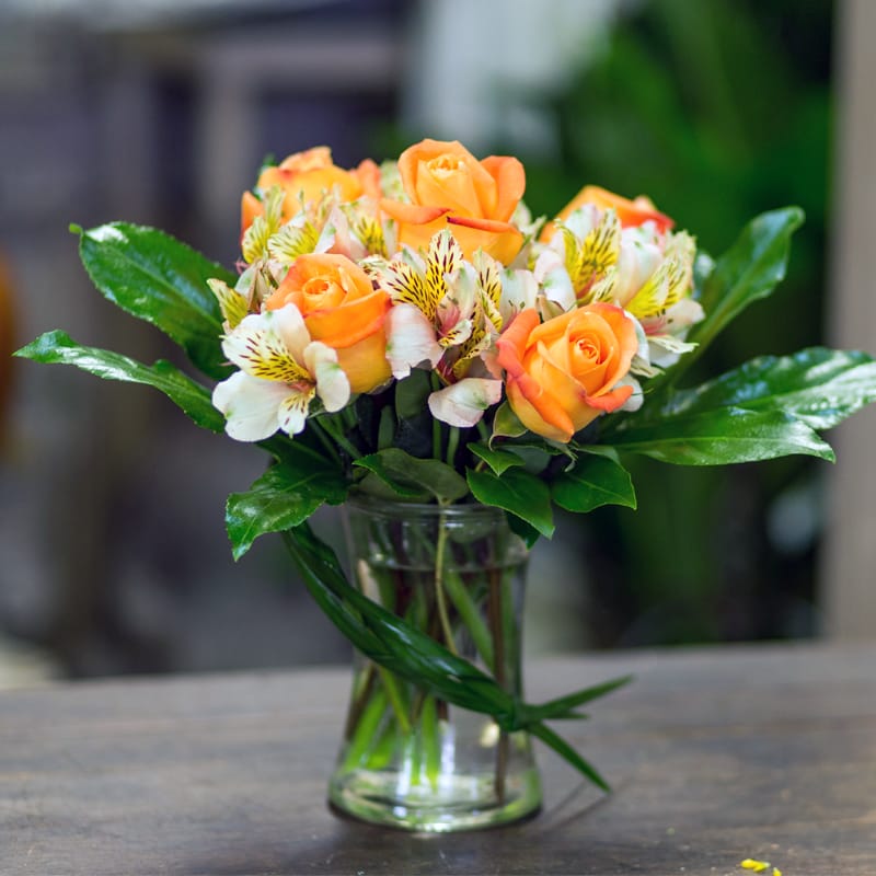 Orange nectar - Bright orange roses and white alstroemeria lilies are grouped together in a clear cylinder vase cuffed with mixed greenery for this eye catching arrangement.