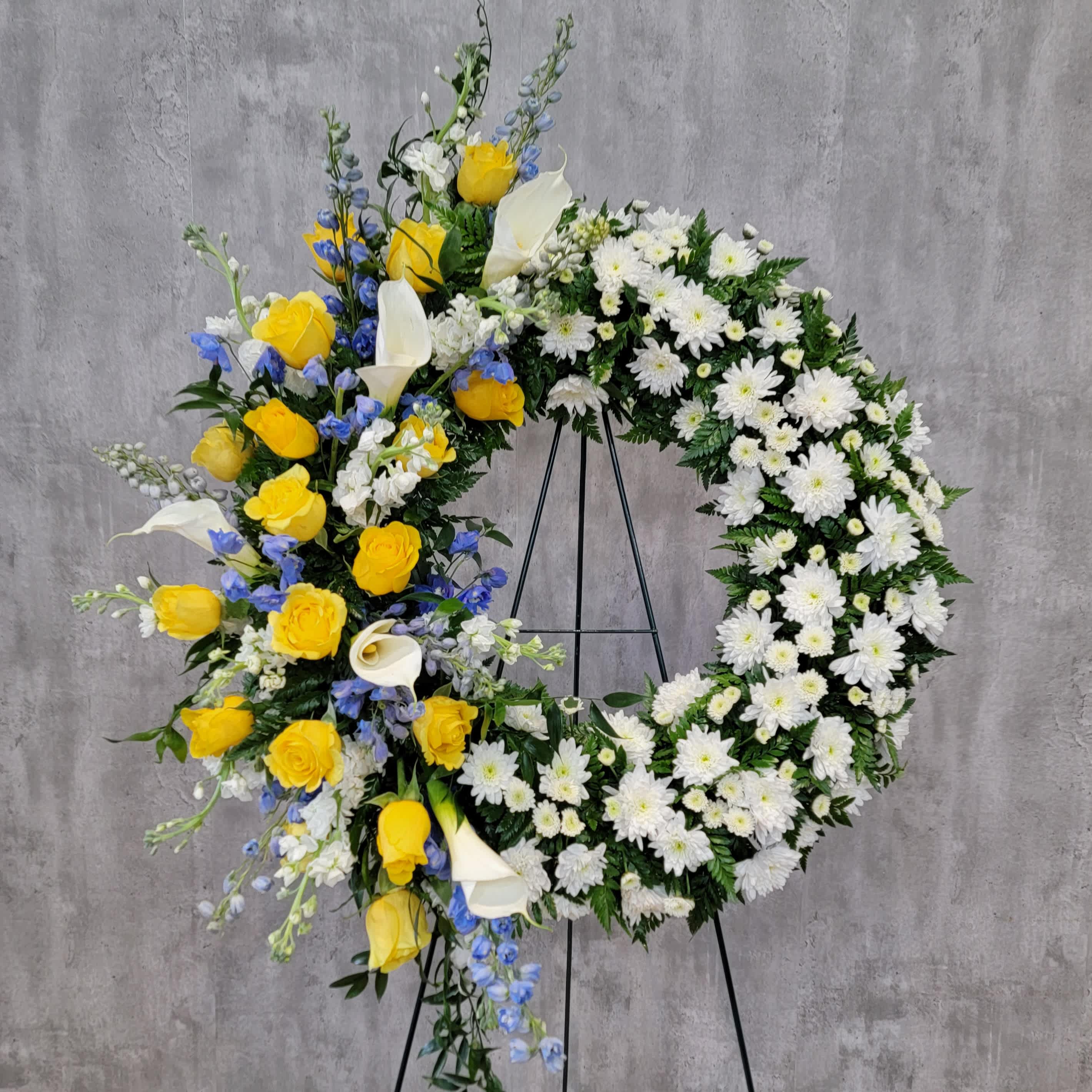 Sunshine Sky Wreath - This beautiful wreath combines the bright colors of yellow and blue against a backdrop of white. The wreath measures approximately 28&quot; across with a swag of calla lilies, yellow roses and blue delphiniums.