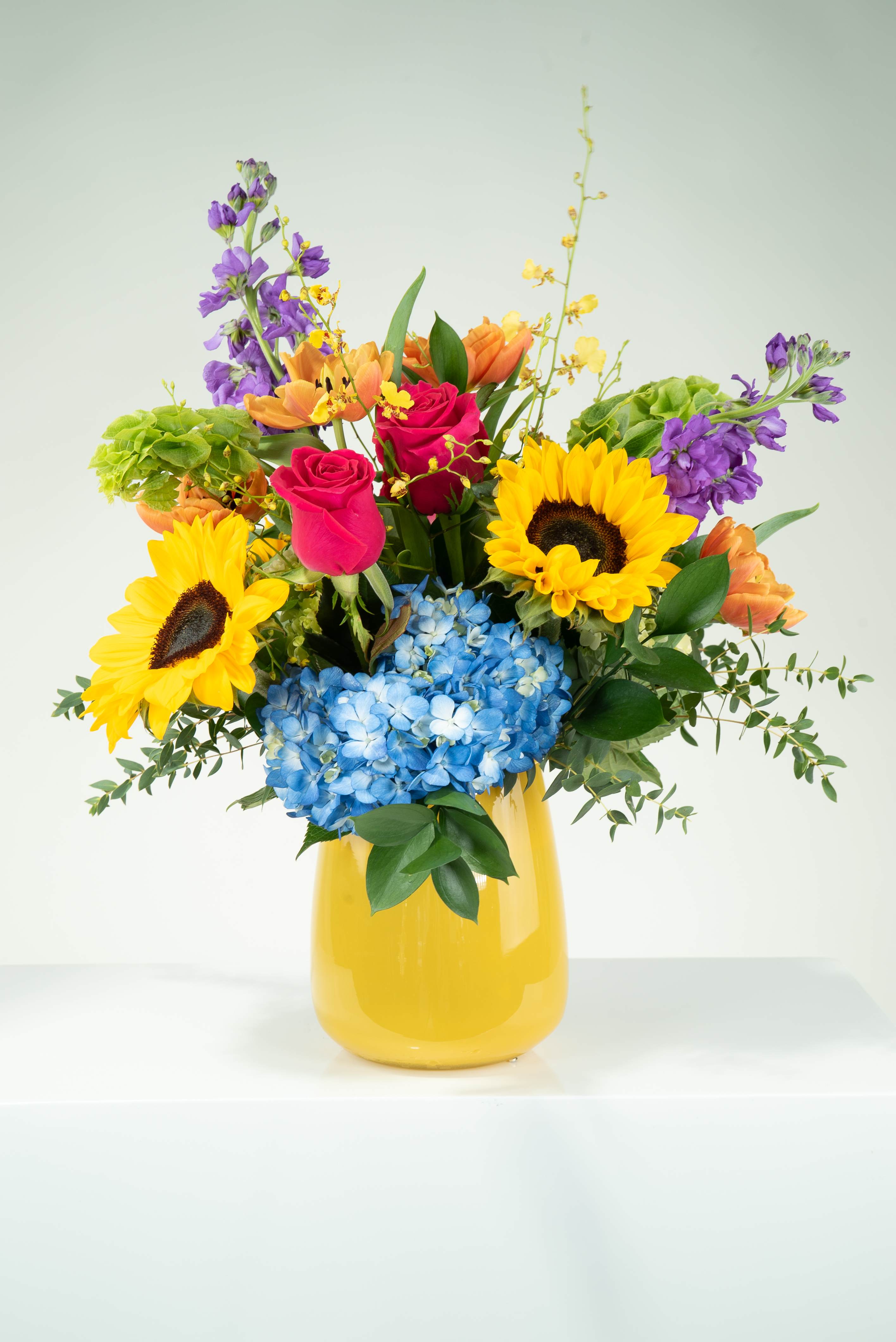 Spring Ahead  - No one is a fan of a time change that makes you lose an hour of sleep. So, we set out to design something nice to look at to make up for the loss. Introducing Spring Ahead: sunflowers, hydrangea, tulips, stock, bells of Ireland, roses, and orchids; complimented with our hand blown polish glass. 