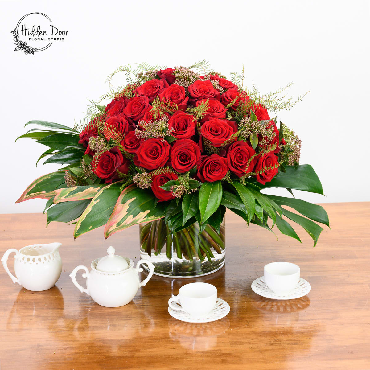 Crimson -  Unlike the ordinary, this arrangement takes these beautiful red roses to a whole new level, showcasing a captivating display of crimson-colored roses and carefully selected seasonal greens. Prepare to be enchanted by the contemporary floral design techniques, which infuse modern flair into this classic arrangement.  &quot;Crimson&quot; is far from your usual bouquet of red roses accompanied by baby's breath. In fact, we do not include any baby's breath in our designs. This arrangement represents a modern interpretation of the classic gesture, expertly showcasing two dozen bright red roses that exude an air of sophistication and luxury. The composition is thoughtfully arranged to create a striking visual impact that is both contemporary and refined. Surrounding the clear vase, you'll find lush green leaves that elegantly support the centerpiece, creating a floating effect atop the vibrant greens. This unique arrangement incorporates bold red roses and various carefully selected seasonal greens, adding depth and texture to the bouquet.   