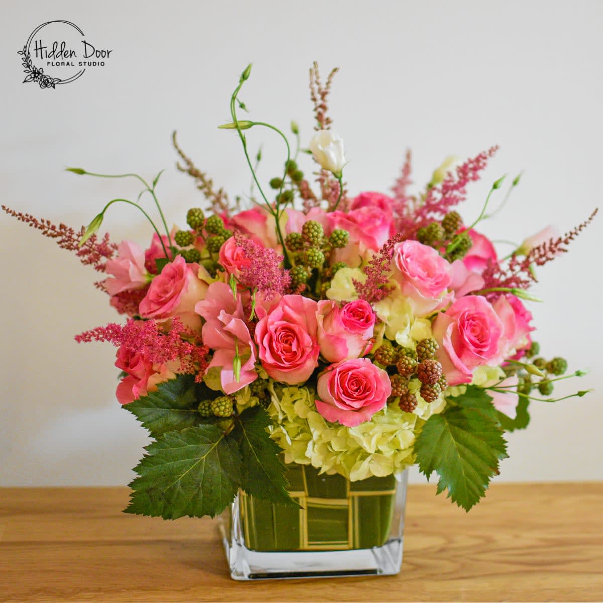 Perfect Harmony Perfect Harmony - The Perfect Harmony arrangement is showcased in a square clear glass vase, adding a touch of contemporary elegance to its romantic appeal. Whether gracing a dining table, adorning a mantle, or serving as a captivating centerpiece, this arrangement effortlessly becomes the focal point, captivating all who look at it. With its romantic look and exquisite combination of blooms, the Perfect Harmony arrangement radiates beauty and sophistication. Its delicate pink roses exude a sense of grace and admiration, while the lisianthus adds an ethereal touch of enchantment. The voluminous hydrangeas create a lush backdrop, further enhancing the arrangement's visual impact. The playful addition of seasonal berries adds a whimsical flair, completing the perfect balance of elegance and charm. At Hidden Door Floral Studio, we understand the importance of creating a memorable and romantic ambiance. The Perfect Harmony arrangement is meticulously crafted to evoke emotions and create lasting impressions. It serves as a testament to our commitment to providing unique and captivating floral designs that capture the essence of every occasion.