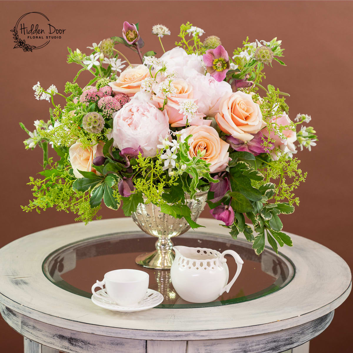 Windsor -   Occasanally when the Peony is off season we will substitute with Garden Rose. This exquisite arrangement, meticulously crafted by the talented artisans at Hidden Door Floral Studio, showcases a stunning combination of peonies, roses, and seasonal flowers, beautifully complemented by accents of lush greenery. Each Windsor arrangement is a work of art meticulously designed to create a dreamy and captivating aesthetic.  The Windsor arrangement is perfect for a variety of occasions and settings. Whether you're planning a grand wedding reception and need stunning centerpieces, organizing a corporate event that calls for an air of sophistication, or simply want to make a romantic gesture to a loved one, the Windsor arrangement will exceed your expectations.  Every Windsor arrangement is uniquely tailored to your preferences and the freshest seasonal blooms available. Our talented floral designers carefully select the finest flowers, ensuring each arrangement is a one-of-a-kind masterpiece. The combination of fragrant peonies, elegant roses, and complementary seasonal flowers creates a harmonious blend of colors, textures, and scents that captivate the senses and leave a lasting impression.       