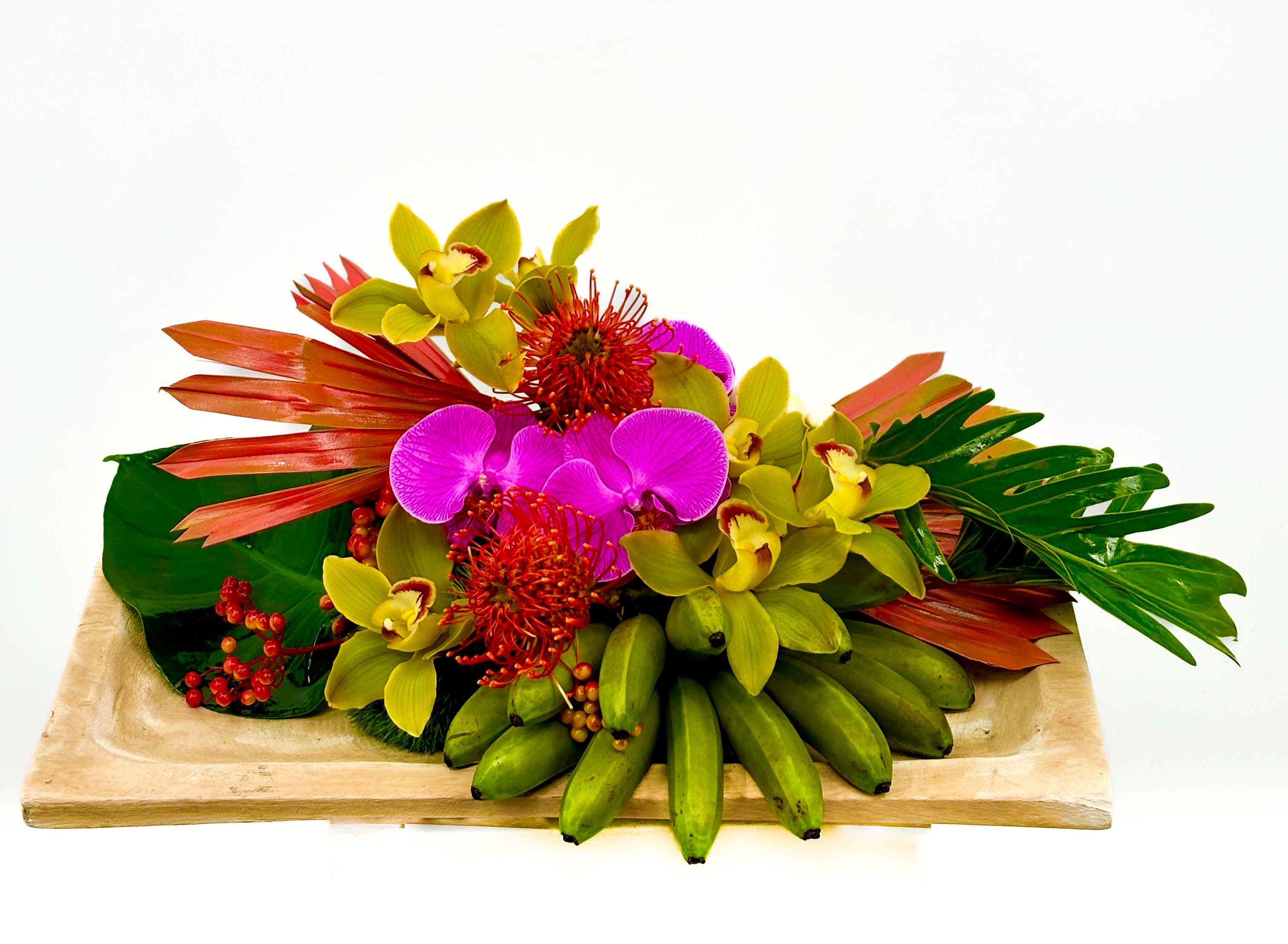 Kalea Kalea - This vibrant floral arrangement features a stunning combination of exotic flowers and lush greenery. Dominating the composition are bright pink orchids, surrounded by yellow-green cymbidium orchids. Adding a splash of fiery color are the red pincushion proteas and red berries, complemented by the striking reddish-orange palm fronds and lush green tropical leaves. The arrangement is accented with a bunch of small green bananas, giving it a tropical and exotic feel. All elements are artfully arranged on a rectangular wooden tray, creating a captivating centerpiece. (Includes 7x14 wood tray)