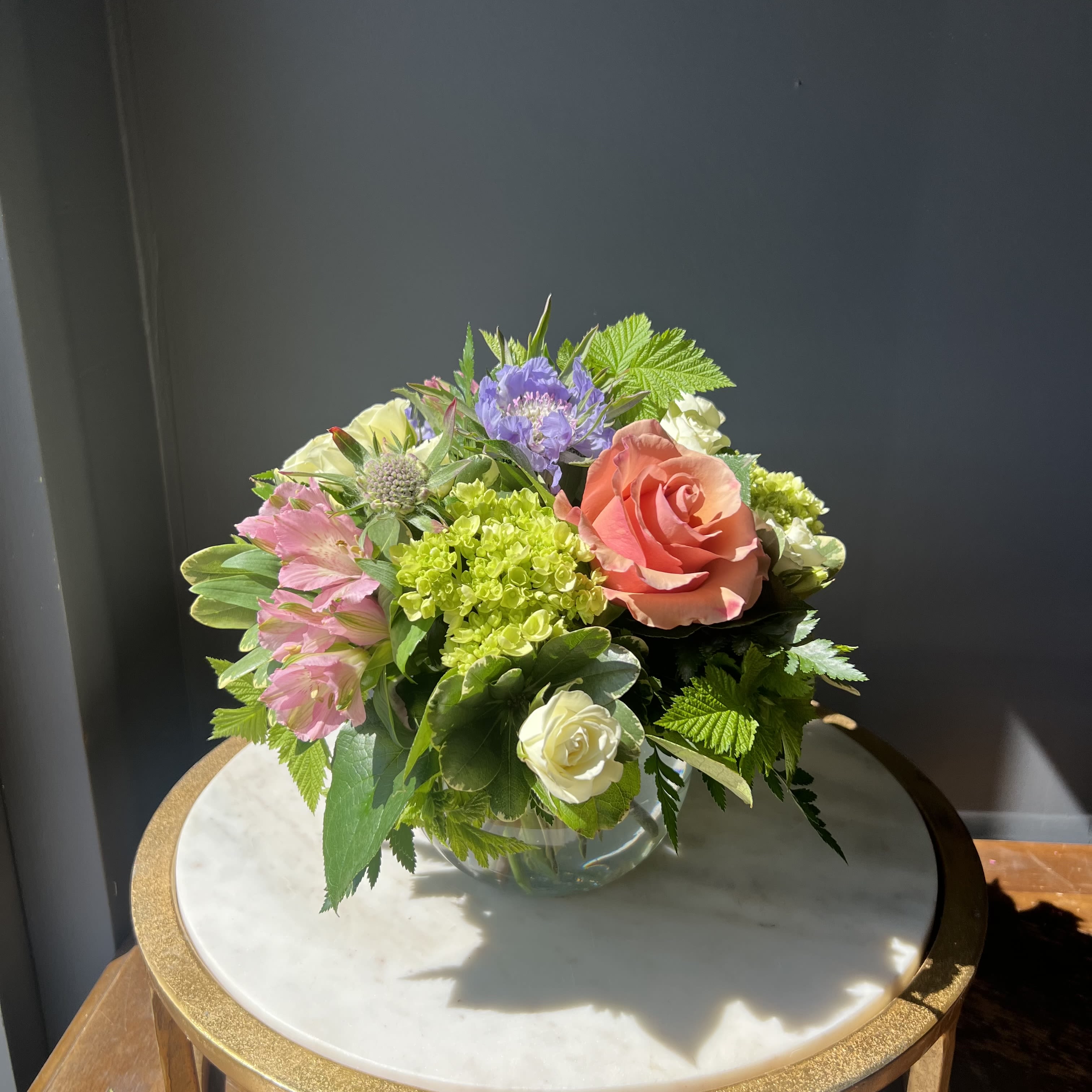 Garden Party - A beautiful spring arrangement in a glass bowl including mini green hydrangeas, roses, and various purple and pink florals.