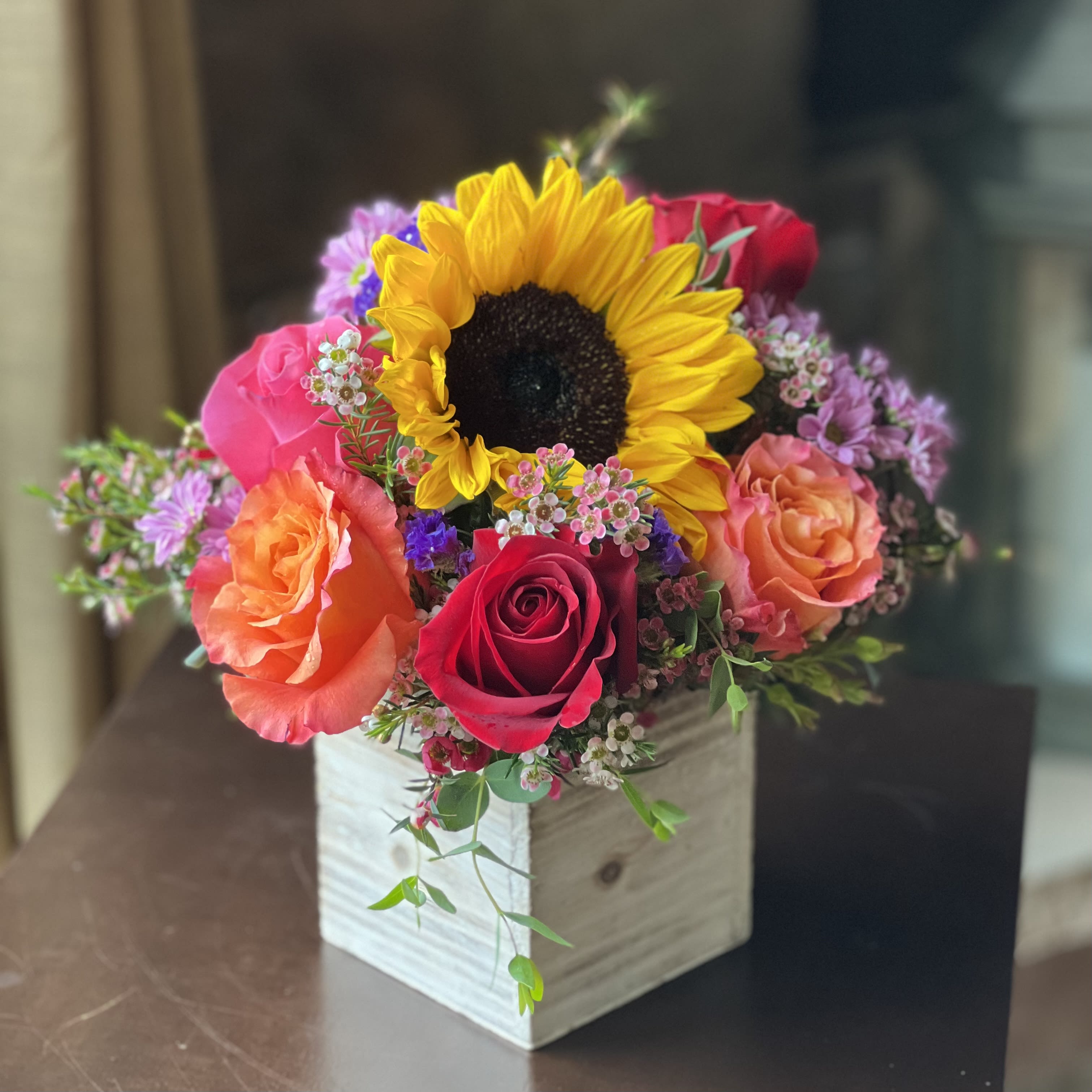 Bursting With Beauty - A whole lot of pretty in a tidy package. This bouquet fits perfectly on a nightstand, a kitchen counter, side-table, or office desk. It will bring a warm glow to any space it fills. Treat someone else or treat yourself to this colorful bouquet!   A great choice for a colorful floral accent on a desk, countertop or bedside table. This arrangement has a rustic wooden container with an inner plastic liner. It should be placed onto a coaster if it is displayed on a moisture sensitive location.  *Please remember that the photos depict a sample arrangement in an overall design style. There are no guarantees that your gift arrangement will look identical to these photos. Tama will do her very best to duplicate the arrangement exactly.