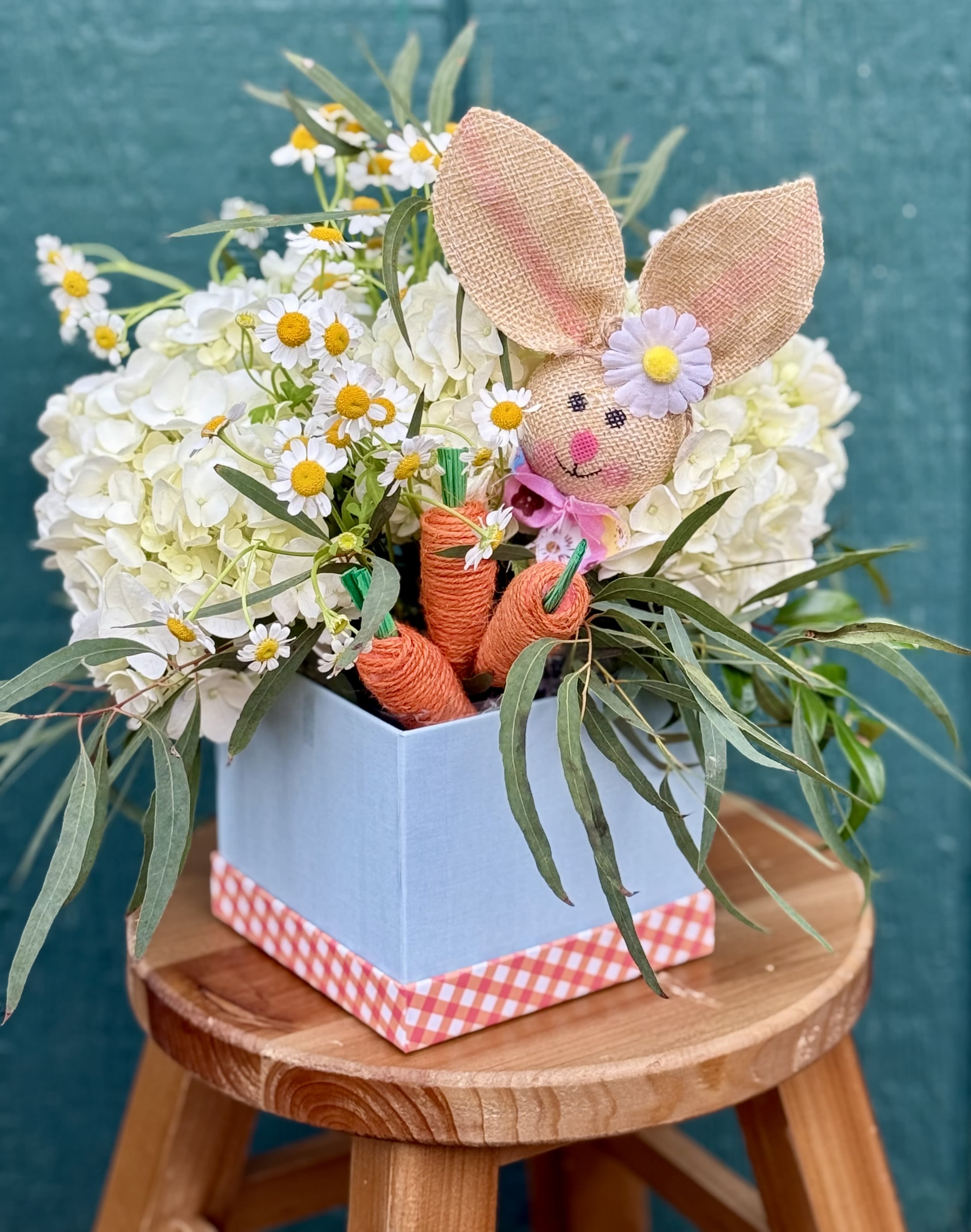 Hoppy Days - A charming nod to springtime delight, this whimsical arrangement features fluffy white hydrangeas and cheerful chamomile blooms nestled among lush greenery. A burlap bunny with rosy cheeks peeks playfully from the garden, surrounded by twine wrapped carrots and gentle pastel touches.  