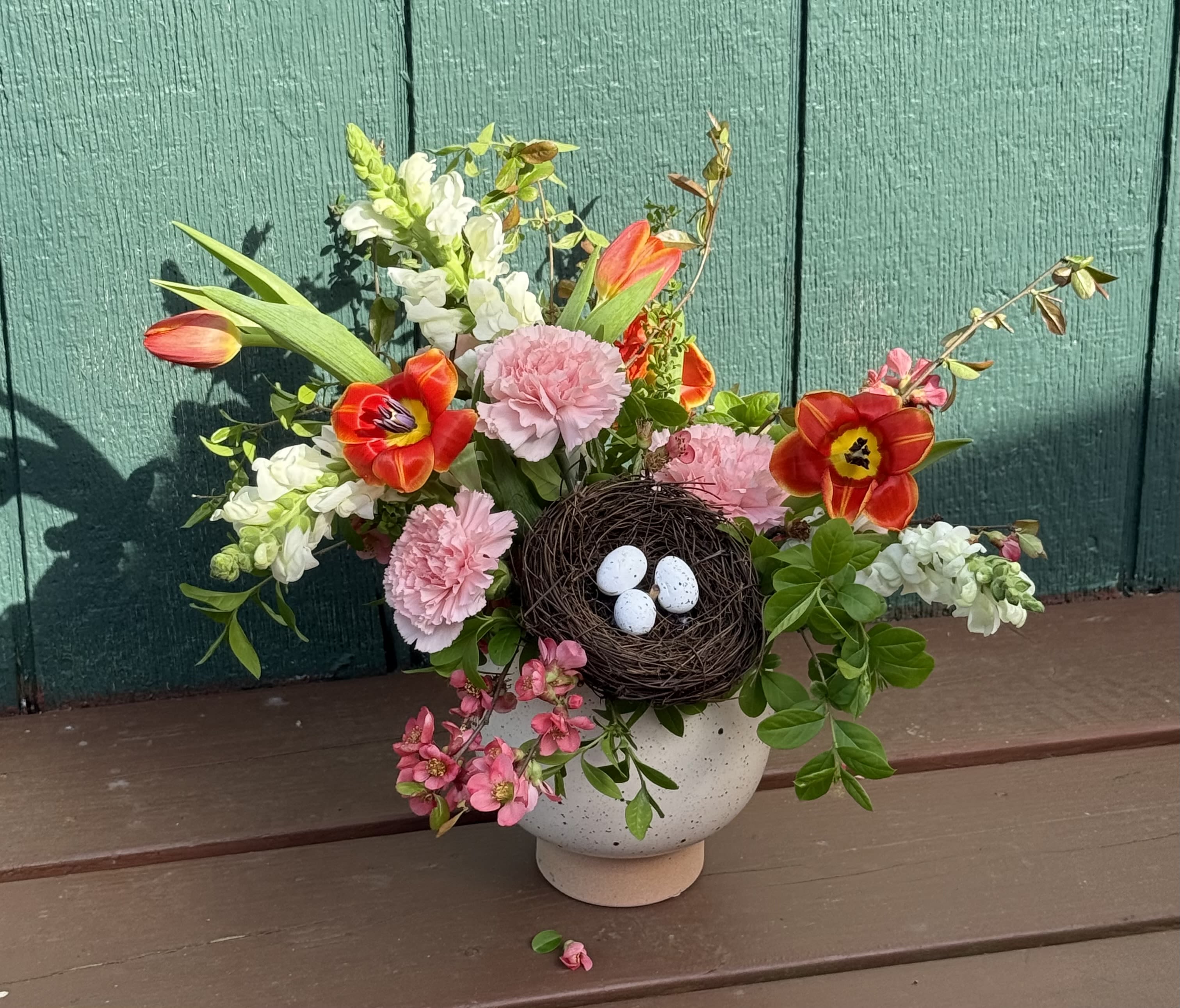 Cheer-y On Top Cheer-y On Top - A celebration of new beginnings, this enchanting garden-style arrangement features vibrant tulips, ruffled carnations, fragrant snapdragons, and delicate spring blooms in shades of coral, blush, and ivory. Nestled at the heart is a charming bird's nest cradling speckled eggs-symbolizing renewal and hope. Designed in a speckled ceramic vase, this piece brings a touch of wild beauty and warmth, perfect for spring gatherings or as a thoughtful seasonal gift.