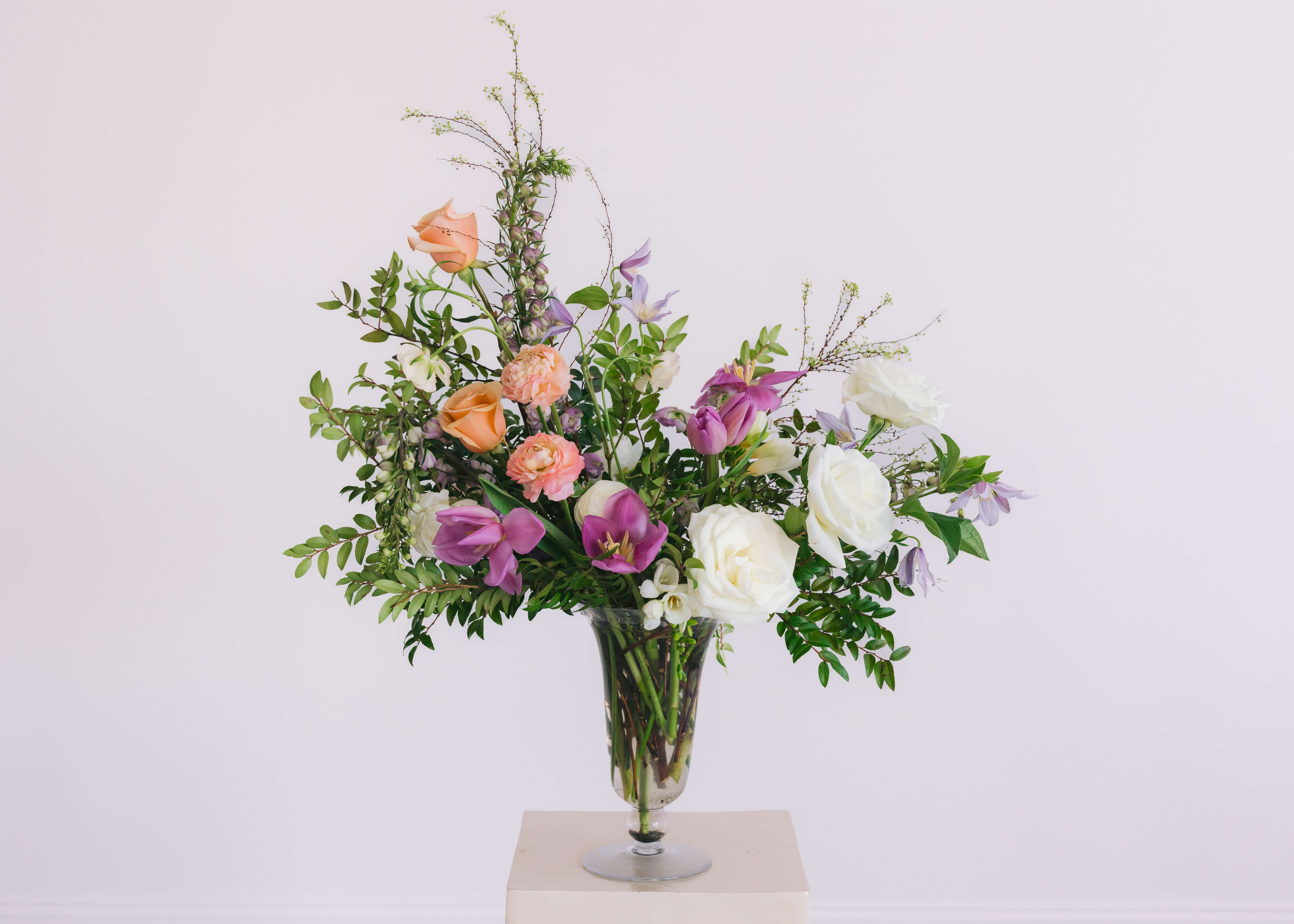 THE CAMBRIDGE THE CAMBRIDGE - A true showstopper designed in an elegant footed glass vase. A larger display of blooms and foliage; with a shape that lends itself to lush, whimsical, garden-style design. Packaged in our signature white tissue and black logo ribbon. Size Pictured: Basic *Please note that our selection of flowers is always changing to ensure a fresh, seasonal mix of blooms. We cannot guarantee specific varieties and we will try to accommodate special requests.