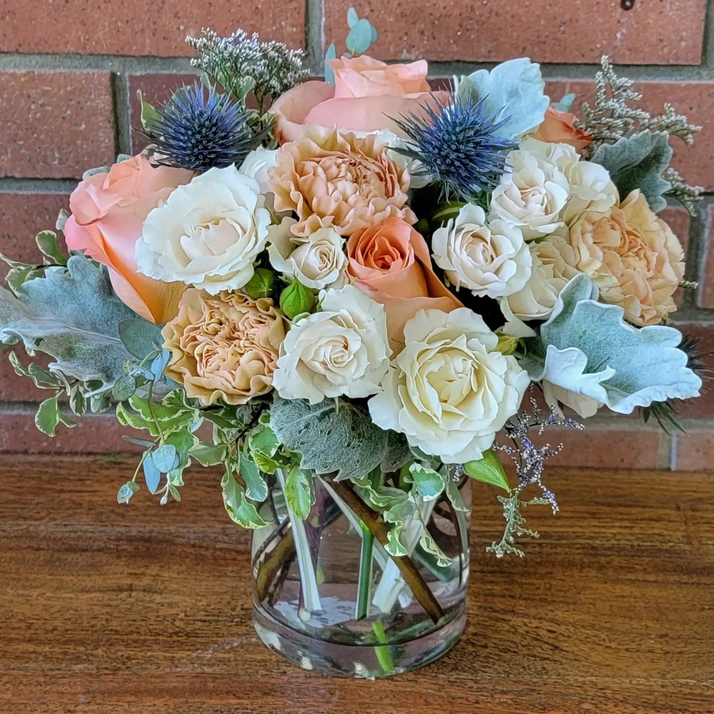 Sweet &amp; Subtle Bouquet - A lovely tabletop fresh flower arrangement with coral roses, blue thistle, Dusty Miller, peachy carnations, creamy white spray roses, greenery and purple whisky filler flower in a heavy glass cylinder vase.
