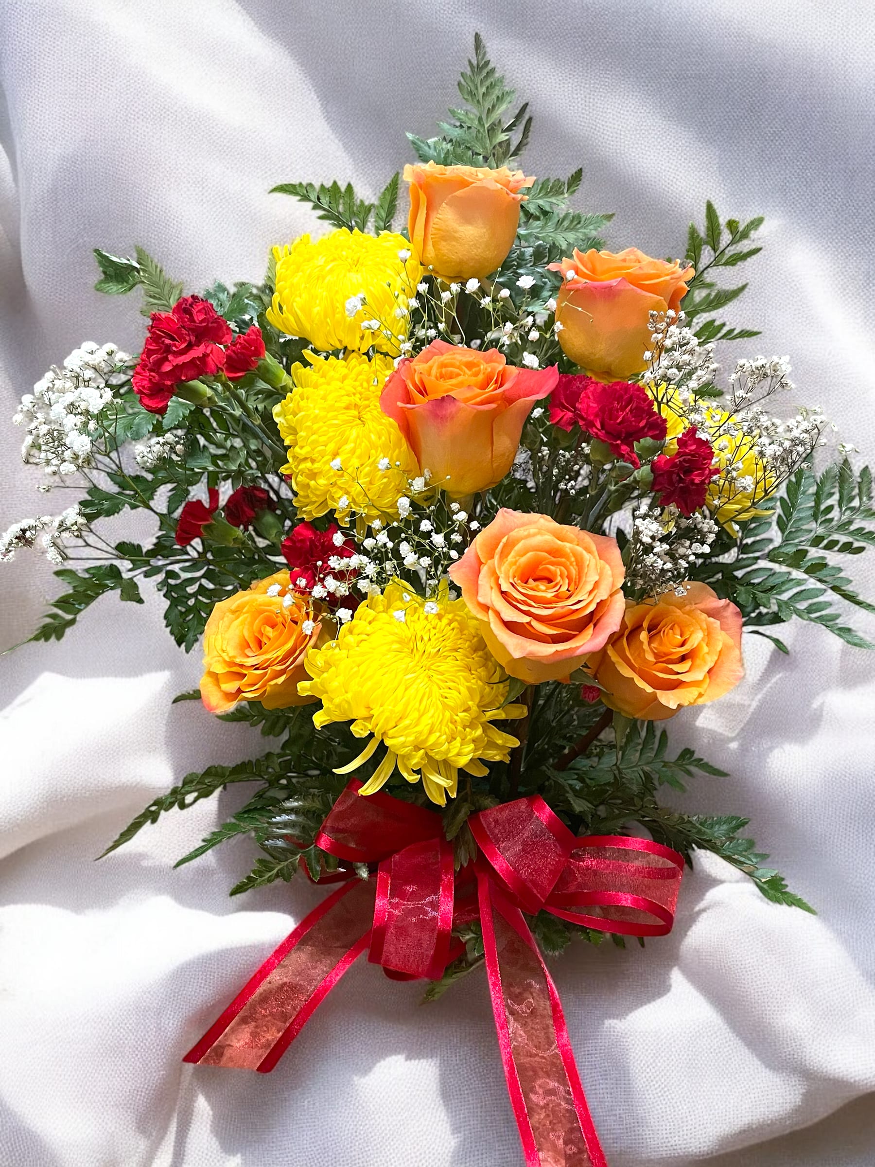 Golden Bloom Golden Bloom - This vibrant floral arrangement features a harmonious blend of colors and textures, exuding warmth and cheerfulness. At its heart are radiant orange roses, their petals glowing with soft, peachy hues. Bright yellow chrysanthemums add a burst of sunshine, perfectly complemented by clusters of deep red carnations that bring a pop of boldness to the mix.