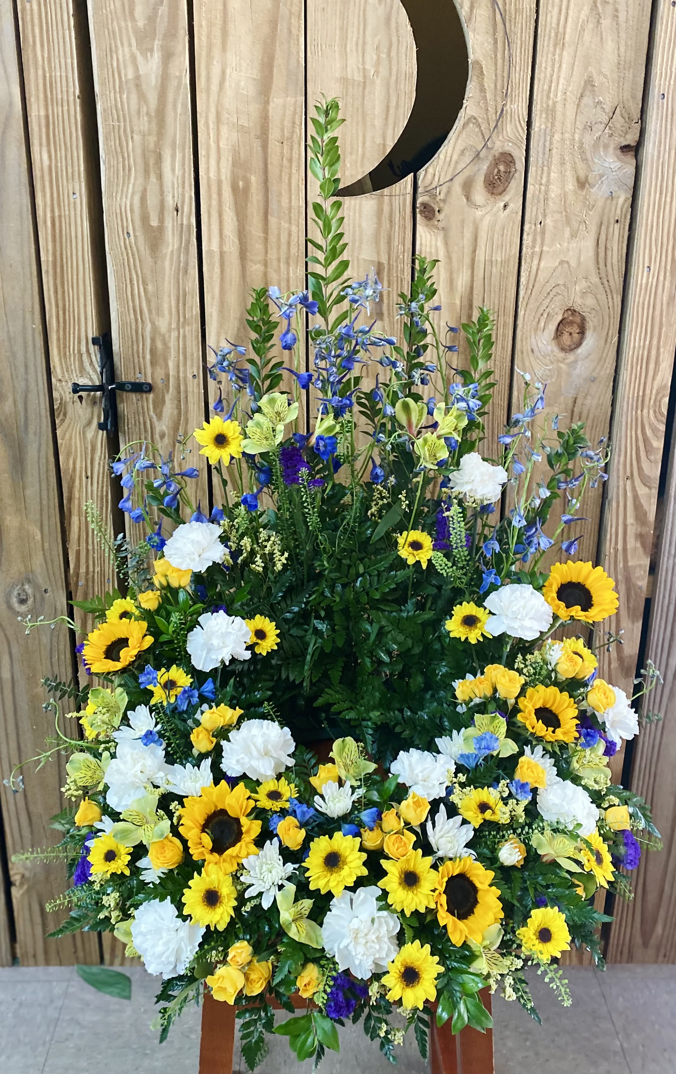 Sunny by the Sea Urn Ring Sunny by the Sea Urn Ring - Sunflowers, blue delphinium, white cushion chrysanthemums, whitecarnations, yellow spray roses, green alstroemeria, and yellow daisies with lepidium to accent this beautiful urn ring.