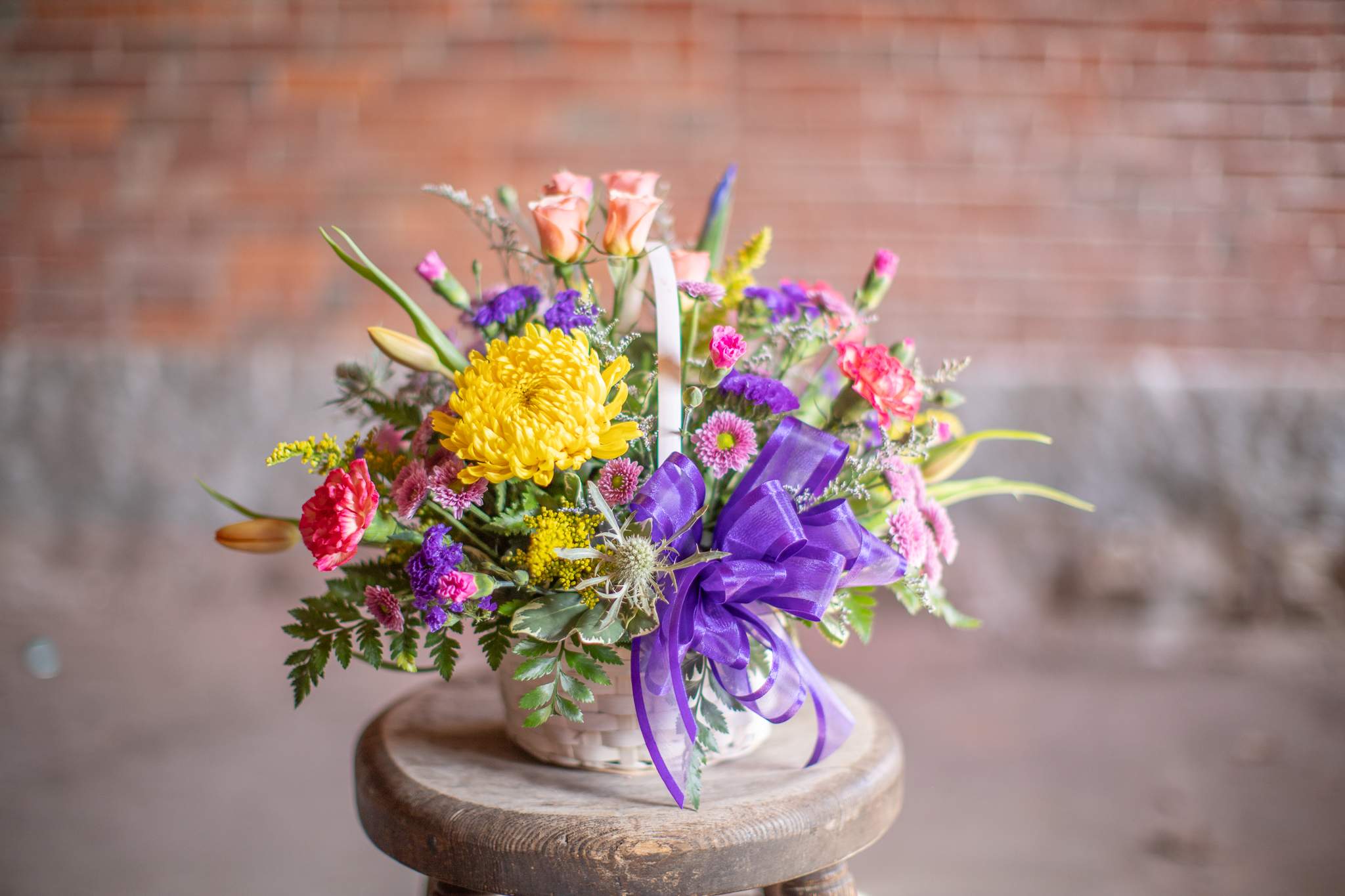Basket of Love - A basket filled with beautiful flowers for your loved one. Pretty cremons, fun button or daisy pon poms and fresh tulips come together harmoniously. Delicate spray roses and carnations complete this one sided arrangement. Upgrades will allow the designer to use more premium blooms like lilies and roses. Basket styles will vary in shape and color tone. Orientation is two sided. This design maybe in a white wash style basket or brown stained wicker. 