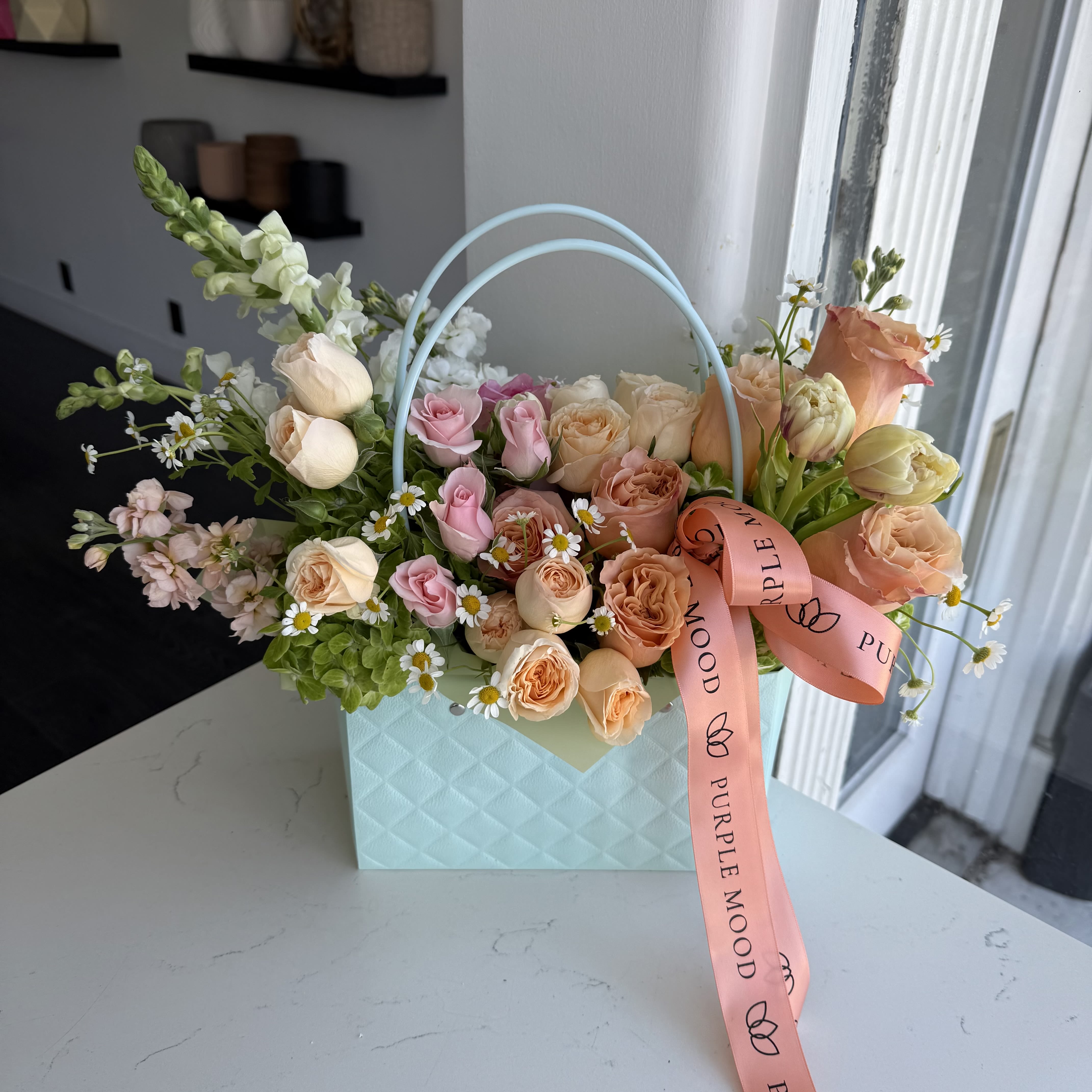 Green Bag Green Bag - A charming floral arrangement presented in a pastel green handbag-style box. This vibrant design features a mix of cheerful roses, bright orange ranunculus, delicate tulips, stock flowers. Perfect for bringing a burst of sunshine to any space, making it an ideal gift for special occasions or a beautiful addition to your home.
