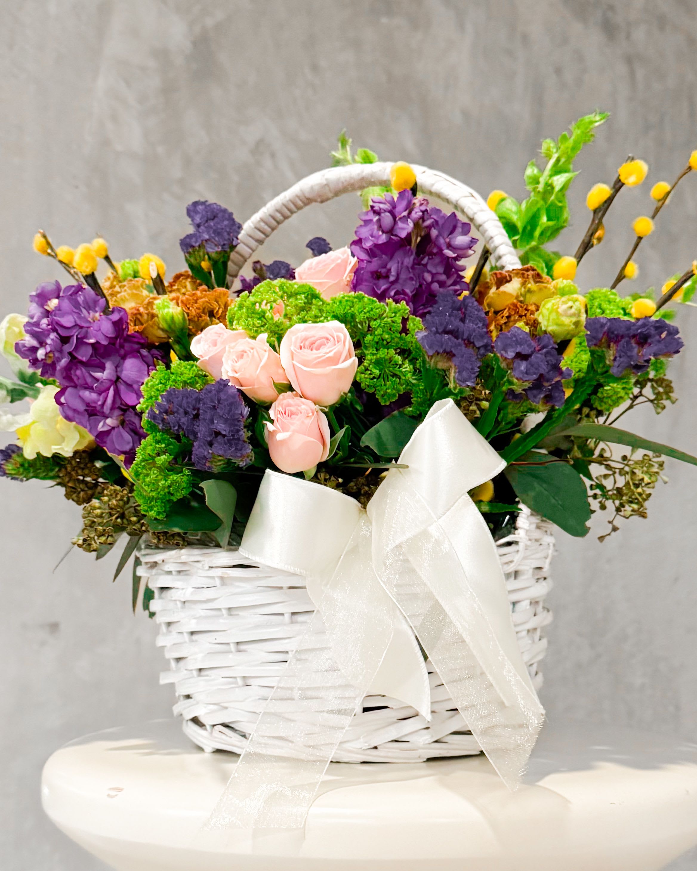 Bounty Hampy - Say hello to Bounty Hampy — a full-on floral moment. This one’s bursting (literally) with bold colors, major texture, and springtime joy. It’s like nature got dressed up for a garden party and brought all her besties. With soft pink roses, juicy purples, sunshiney yellow poms, and those cool green textures, it's giving floral maximalism in the best way. Wrapped up in a crisp white basket with a sheer bow, it’s ready to turn any day into a celebration. 