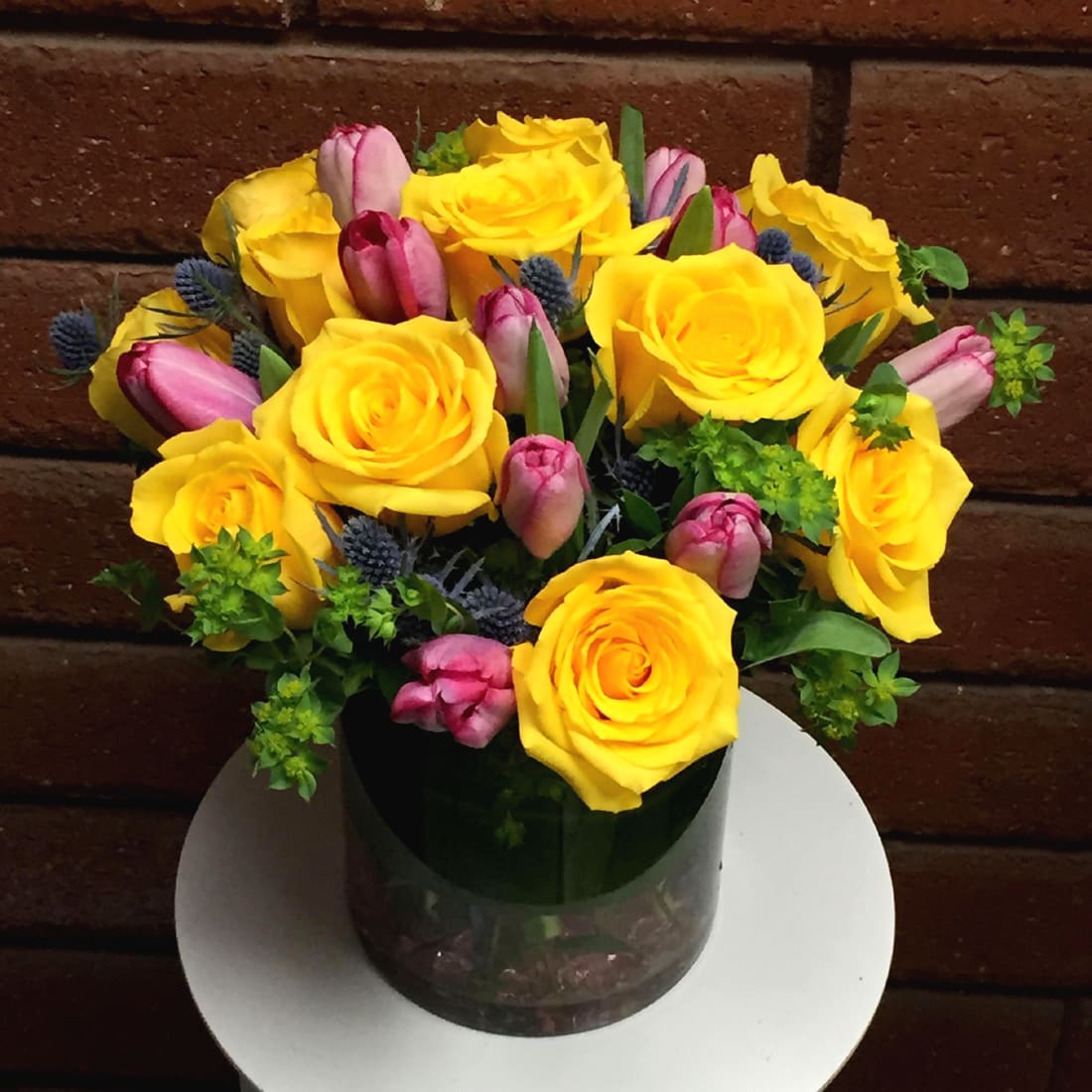 Yellow Roses and Pink Tulips Vase - A burst of exquisite color, House of Stemms delivers yellow roses and pink tulips, accented with blue thistle and fresh garden greens. Bouquet is hand arranged and delivered in a clear glass vase, lined with a tropical leaf and colored beads. Standard size is approximately 10in (W) x 14in (H).  Standard - 12 Yellow Roses, 10 Pink Tulips, Blue Thistle and Garden Greens - 6in (W) Cylinder Vase  Deluxe - 18 Yellow Roses, 15 Pink Tulips, Blue Thistle and Garden Greens - 6in (W) Cylinder Vase  Premium - 24 Yellow Roses, 20 Pink Tulips, Blue Thistle and Garden Greens - 8in (W) Cylinder Vase  Care Tips: Place your bouquet in a cool location away from direct sunlight, heating or cooling vents, drafty places, directly under ceiling fans, or on top of televisions or radiators. Check water level daily, keep the vase full with clean water. Change water every 2-3 days and apply a sharp fresh cut to the stems to ensure extended flower's life span.