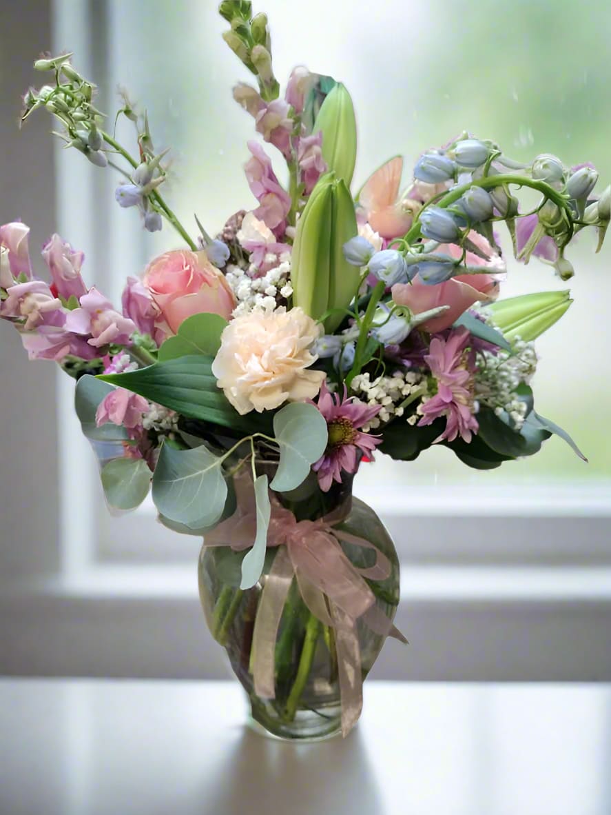 Gentle Hues Vase Arrangement - The Gentle Hues Vase arrangement showcases the softest tones of fresh blooms, harmoniously blended to form a stunning floral display that is ideal for any event or memorial service. This exquisite arrangement may feature roses, delphiniums, carnations, lilies, and snapdragons, all coming together to create a serene and elegant look. When you request custom designs, we will strive to craft arrangements that closely mirror the image you have in mind. However, please remember that each creation is one-of-a-kind and tailored specifically for you. Given the natural variability of flowers, some color substitutions might be necessary, and the prices and availability of seasonal blooms can vary as well.