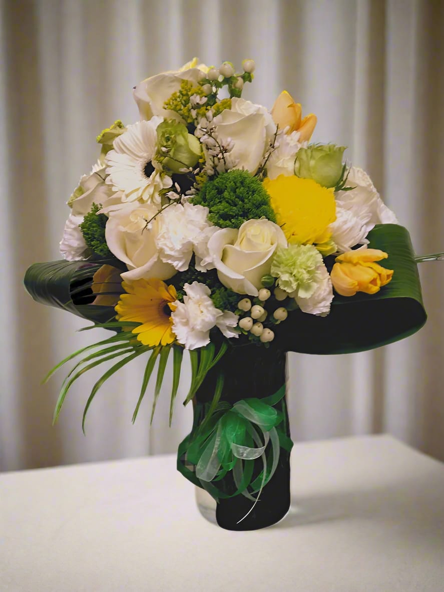 Peeking Sunshine Vase - A stunning vase brimming with vibrant blooms and lush greenery features Gerbera daisies, roses, carnations, hypericum berries, trichillium, and white tulips, all accented by cheerful hints of green and yellow that evoke the warmth of sunshine and the freshness of grass.  **When you request custom designs, we will create them to closely resemble the picture. Keep in mind that each design is unique and made just for you. Due to the nature of flowers, color substitutions may be required. Prices and availability of seasonal flowers can also differ.