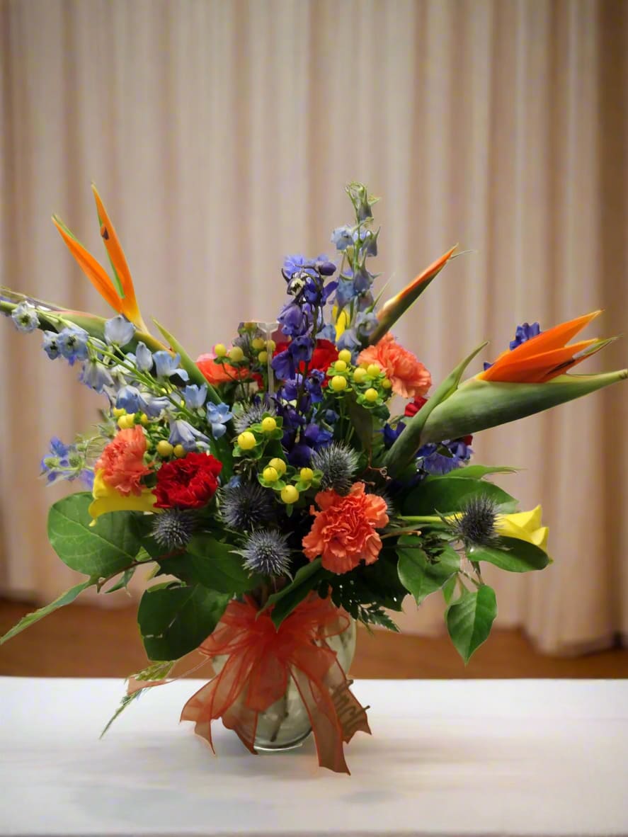 Primary Textures Vase - A stunning display of primary colors and rich textures comes to life in this arrangement. Vibrant delphinium shades blend beautifully with bird of paradise, carnations, globe thistle, and hypericum berries, all elegantly presented in a glass vase adorned with a charming bow.  When you request custom designs, we will create them to closely resemble the picture. Keep in mind that each design is unique and made just for you. Due to the nature of flowers, color substitutions may be required. Prices and availability of seasonal flowers can also differ.