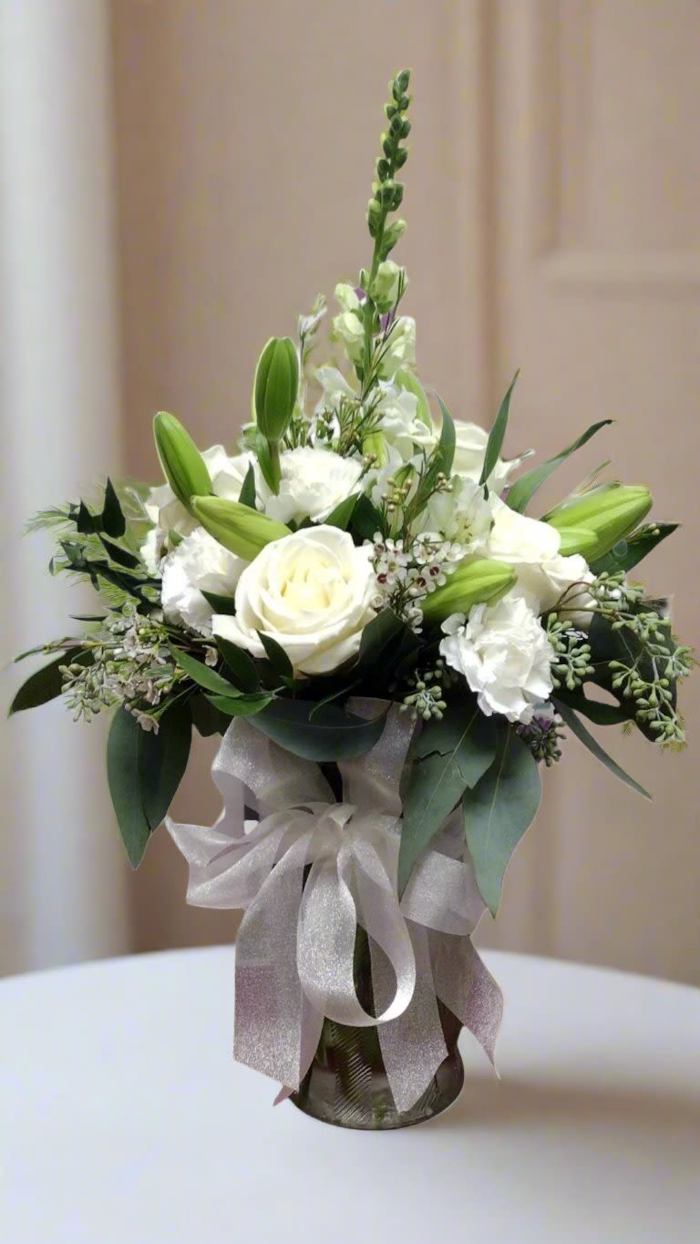 Elegant White Vase - Discover the Elegant White Vase, a chic glass vase bustling with fresh flowers, featuring gorgeous white roses, lilies, snapdragons, and mini carnations, enhanced by fillers and greens, all beautifully adorned with a bow. It's the perfect statement for any occasion.  &nbsp;When you request custom designs, we will create them to closely resemble the picture. Keep in mind that each design is unique and made just for you. Due to the nature of flowers, some substitutions may be required. Prices and availability of seasonal flowers can also differ.
