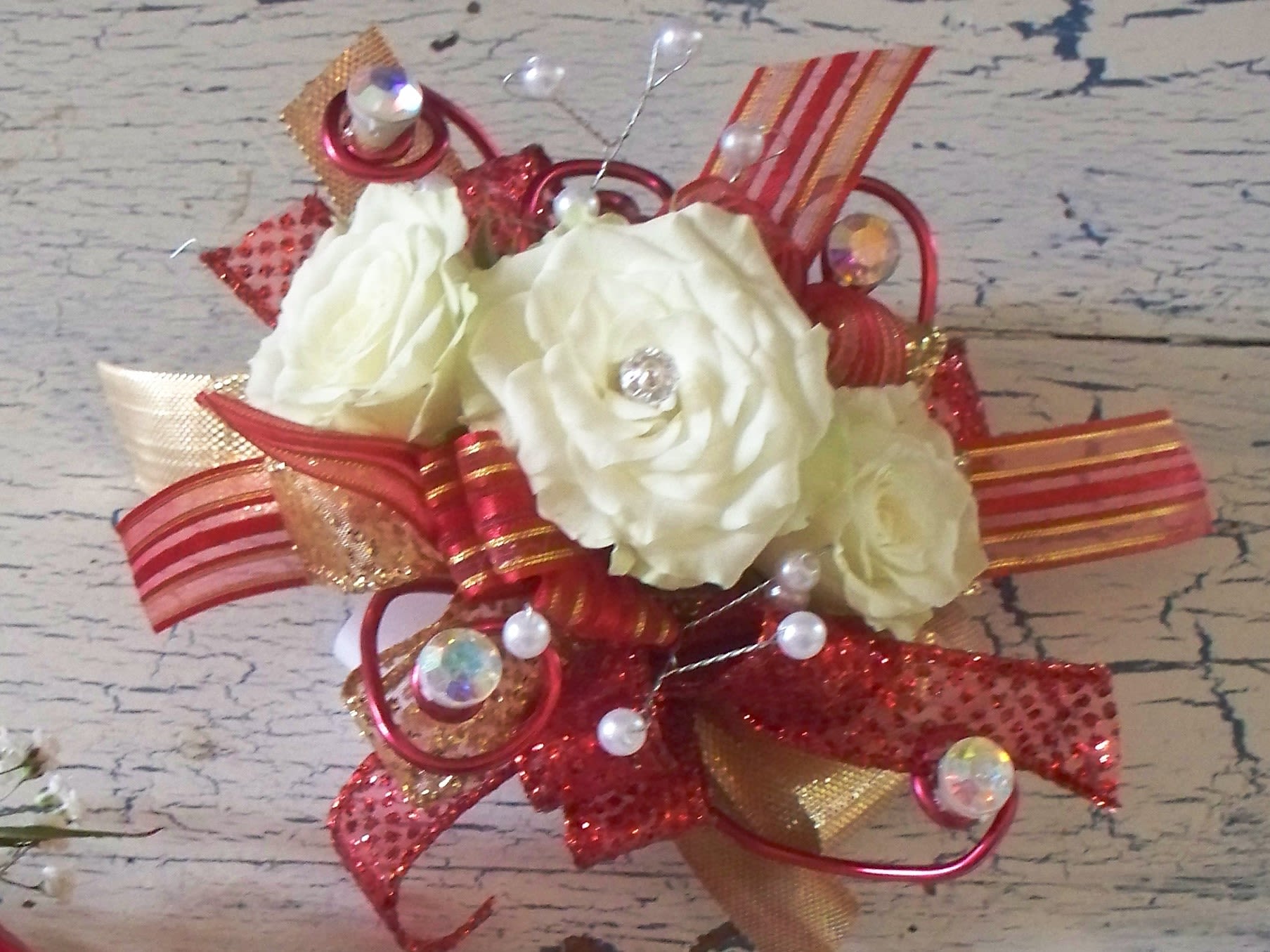 Glitz Wrist Corsage Glitz Wrist Corsage - Gems and pearls, red and gold ribbons with red wire accents & rhinestone accents makes the white rose glitzy. Included is a disposable elastic wristlet, maybe upgrade to a dazzle rhinestone wristlet.