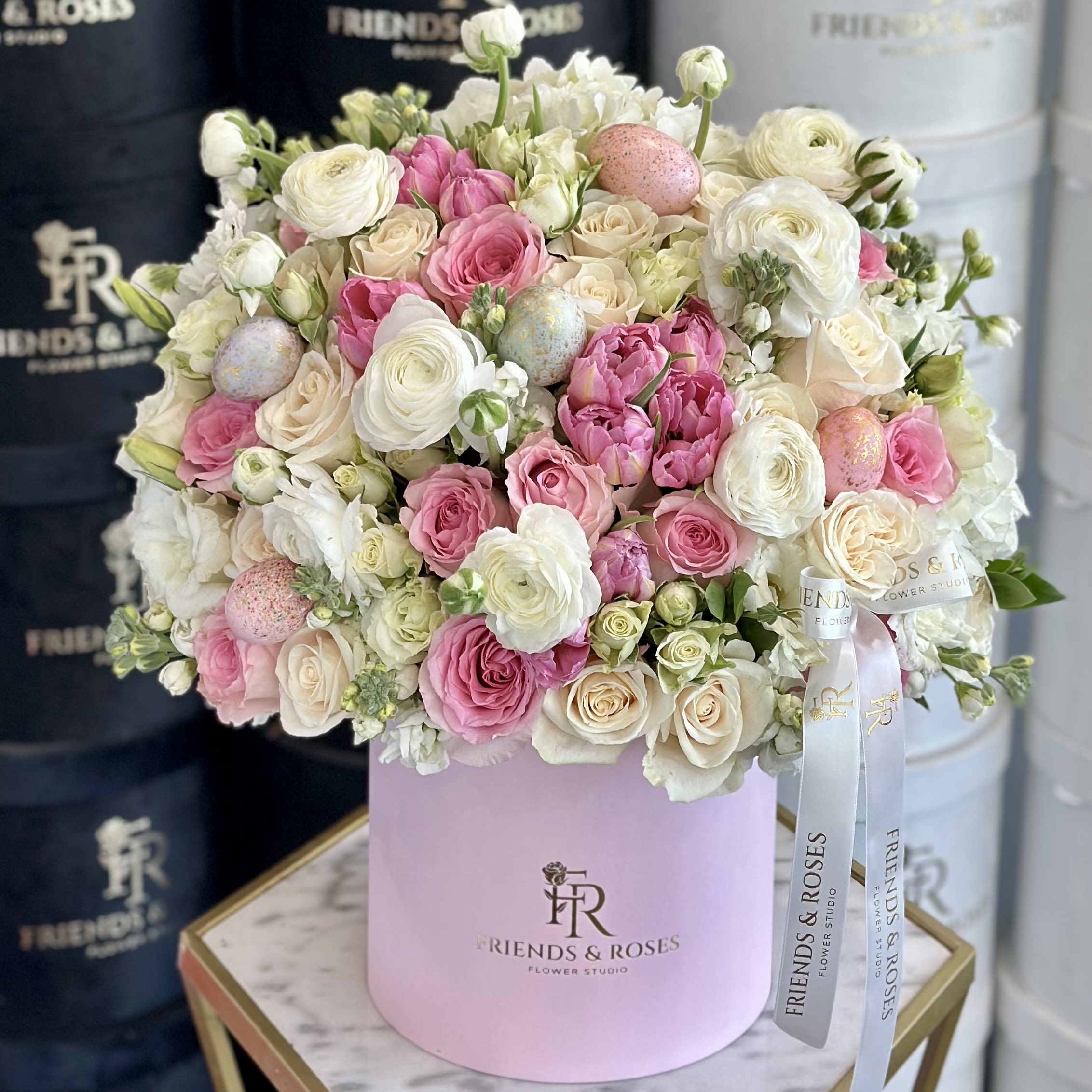 Egg-stra Special Spring Flowers - A soft, pastel dream unfolds, where the elegance of blush white and pink roses, creamy tulips, and vibrant ranunculus dance together in harmony. The flowers are artfully nestled amidst tender greenery, with the final touch of decorative eggs scattered like hidden treasures. This luxurious display celebrates spring's finest, exuding both the purity and joy of the season, and bringing a touch of grace and whimsy to any space.