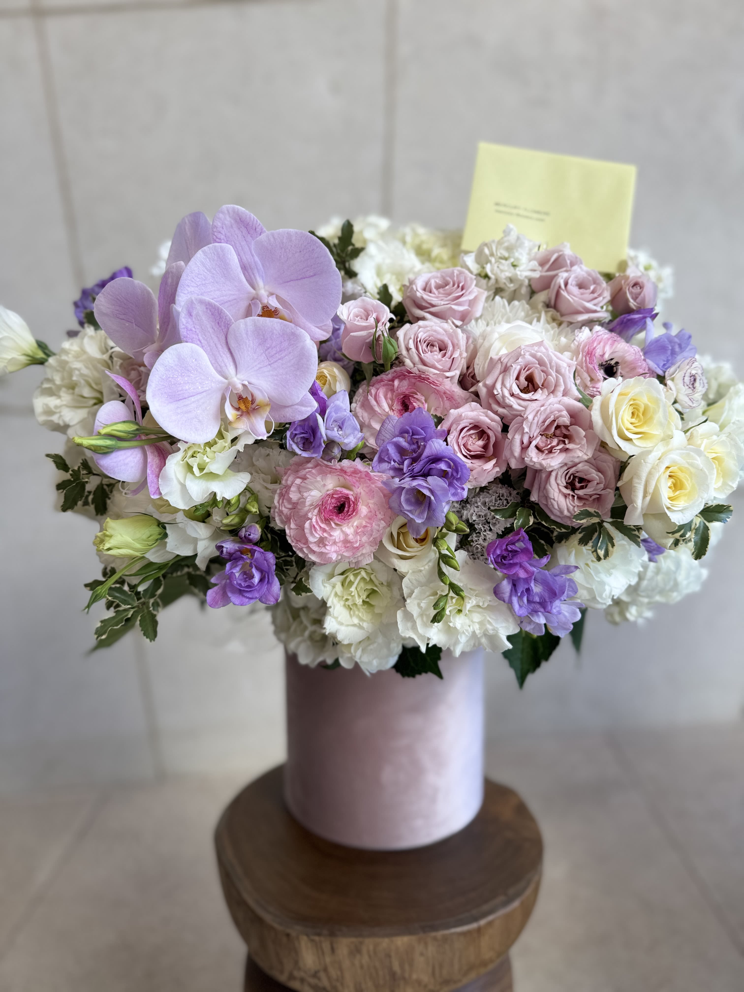 Pastel Candy Box - This is an elegant floral arrangement in soft pastel tones, presented in a luxurious velvet pink box. The composition features delicate pale pink and lavender roses, lush white hydrangeas, and vibrant purple lisianthus, complemented by soft cream and blush accents. Orchid blooms add a touch of exotic beauty, while delicate ranunculus and other seasonal flowers enhance the romantic and sophisticated feel. This charming bouquet exudes grace, softness, and timeless elegance, making it perfect for any special occasion or to simply brighten someone’s day.