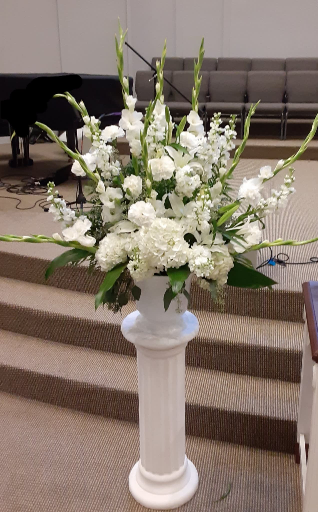 White Wedding Event Floral Urn - An assortment of beautiful white flowers, such as gladiolus, stock, lisianthus. roses and more with assorted greeneries in a white traditional urn for the wedding
