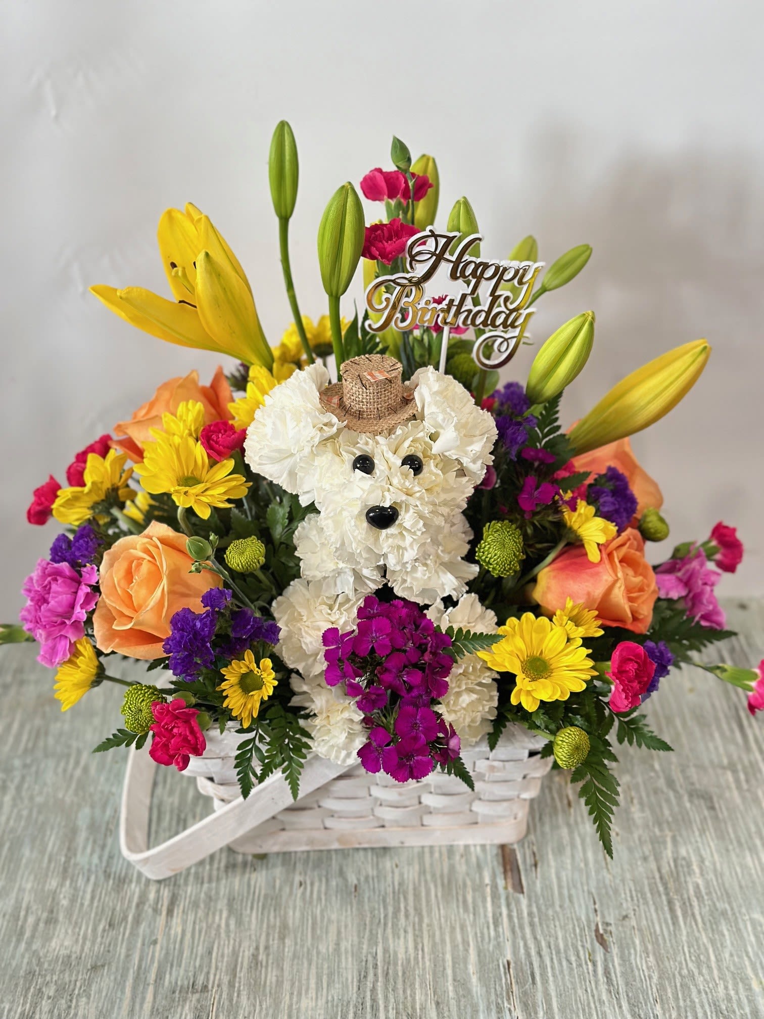 Birthday #12 Birthday #12 - Arrangement of fresh cut flowers that includes a puppy made out of all white carnations and is adorned with a little hat. Designed in a white rectangular basket. Embellished with a gold "happy birthday" pick. Colors can be modified depending on availability.
