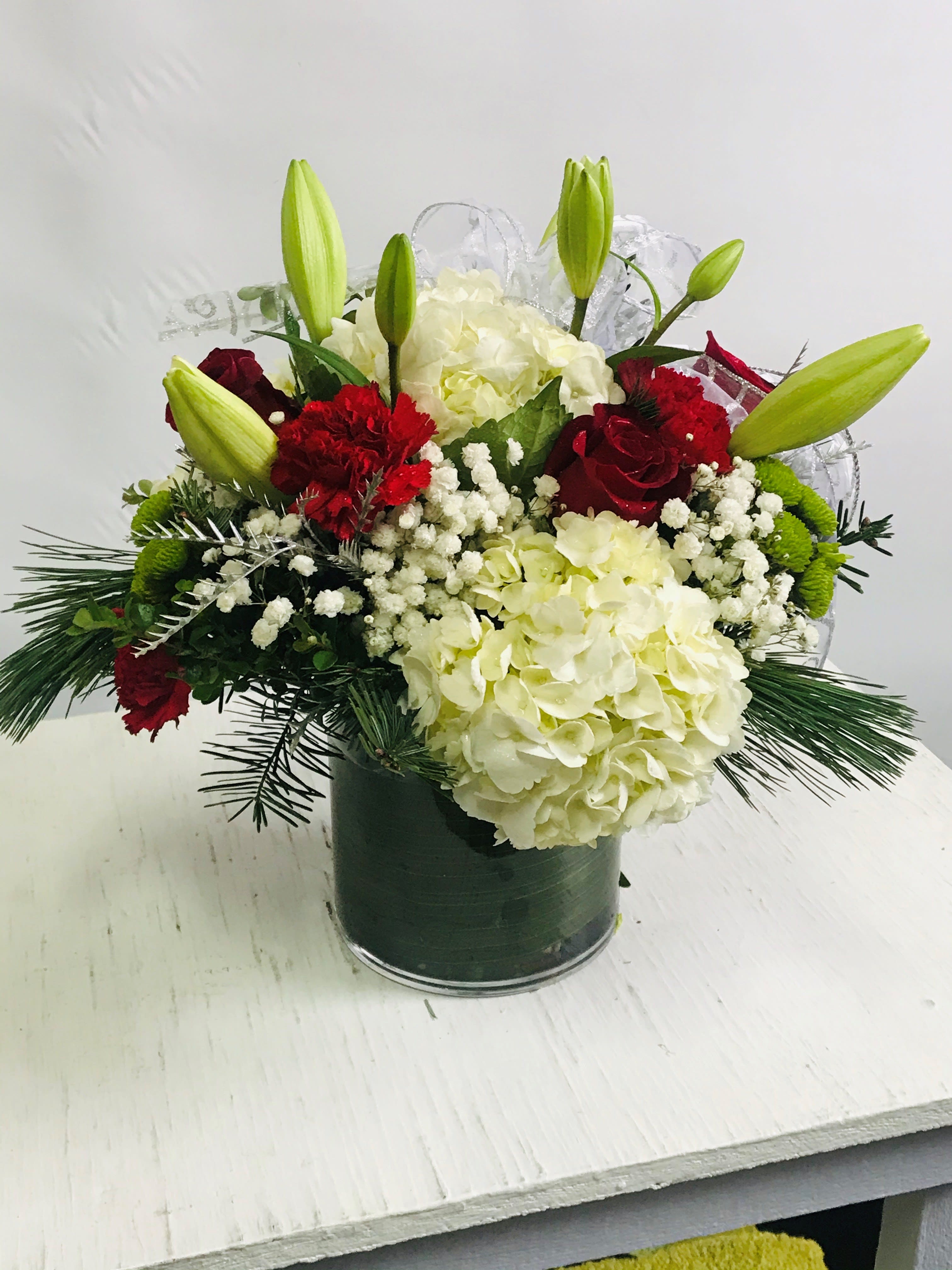 Christmas #6 Christmas #6 - An arrangement of fresh cut flowers designed in a clear glass cylinder vase lined with Ti leaf ribbon. Arrangement to include seasonal Christmas greens.
