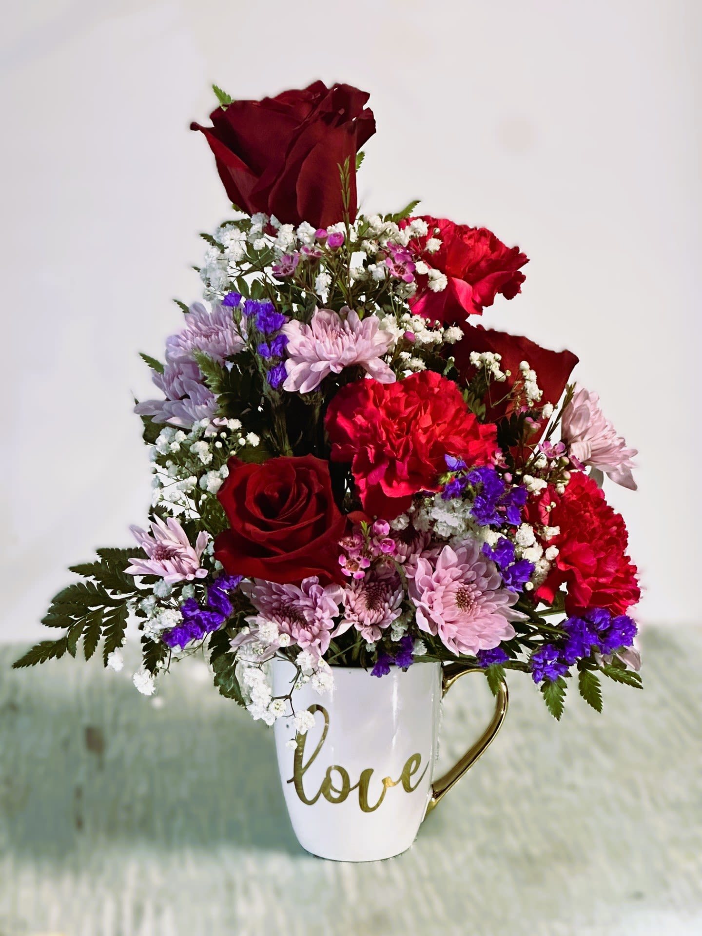 Love and Romance #2 - Arrangement of red roses, magenta carnations, lavender cushion poms, purple statice, babies breath &amp; leather fern arranged in a ceramic mug(12oz.) that reads &quot;Love&quot; in metallic gold print.
