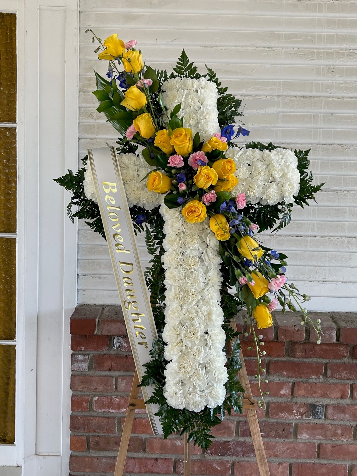 Sympathy Cross #54 - All white standing cross with a cluster of yellow, pink and purple fresh cut flowers. Colors can be modified depending on availability. Designed on an easel that is included with the arrangement and does NOT need to be returned. Banner is included with arrangement. Please specify what is to be printed on banner upon check out under &quot;FLORIST INSTRUCTIONS&quot;. LIMITED TO 5 WORDS ON BANNER.