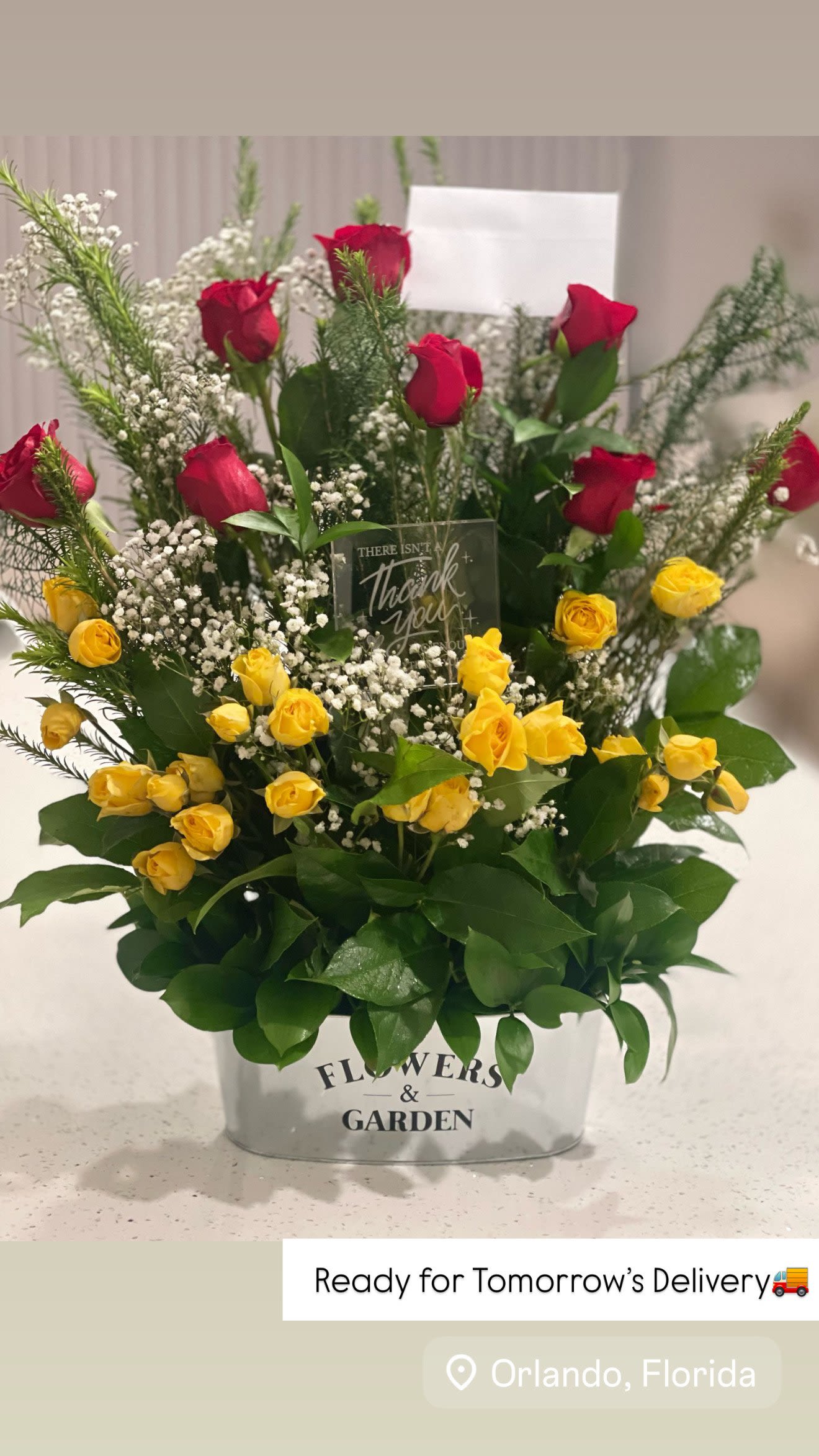 Gratitude in Bloom Arrangement Gratitude in Bloom Arrangement - Send a heartfelt message of appreciation with our stunning “Gratitude in Bloom” floral design. This lush arrangement features a harmonious mix of deep red roses, cheerful yellow spray roses, and delicate baby's breath, beautifully accented with fresh greenery for a vibrant, full look. Nestled in a charming white "Flowers & Garden" tin planter, it's topped with a sweet acrylic sign that reads, “There isn’t a thank you big enough for you.” Perfect for thank-you gifts, celebrations, or simply brightening someone’s day.