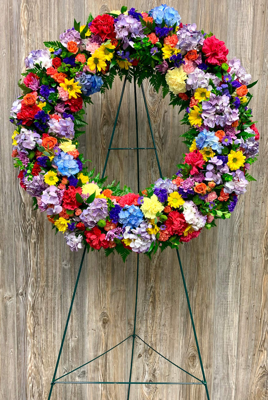 Large Rainbow Wreath Large Rainbow Wreath - This vibrant Standing Easel for Funeral Rainbow Wreath is a beautiful testament to the life of the deceased. The wreath is done in a colorful array of rainbow colors, creating an eye-catching display. Its bold and bright presentation will bring comfort to all in attendance, honoring the life and spirit of your loved one with a stunning and lasting reminder of their life. *Flowers may vary