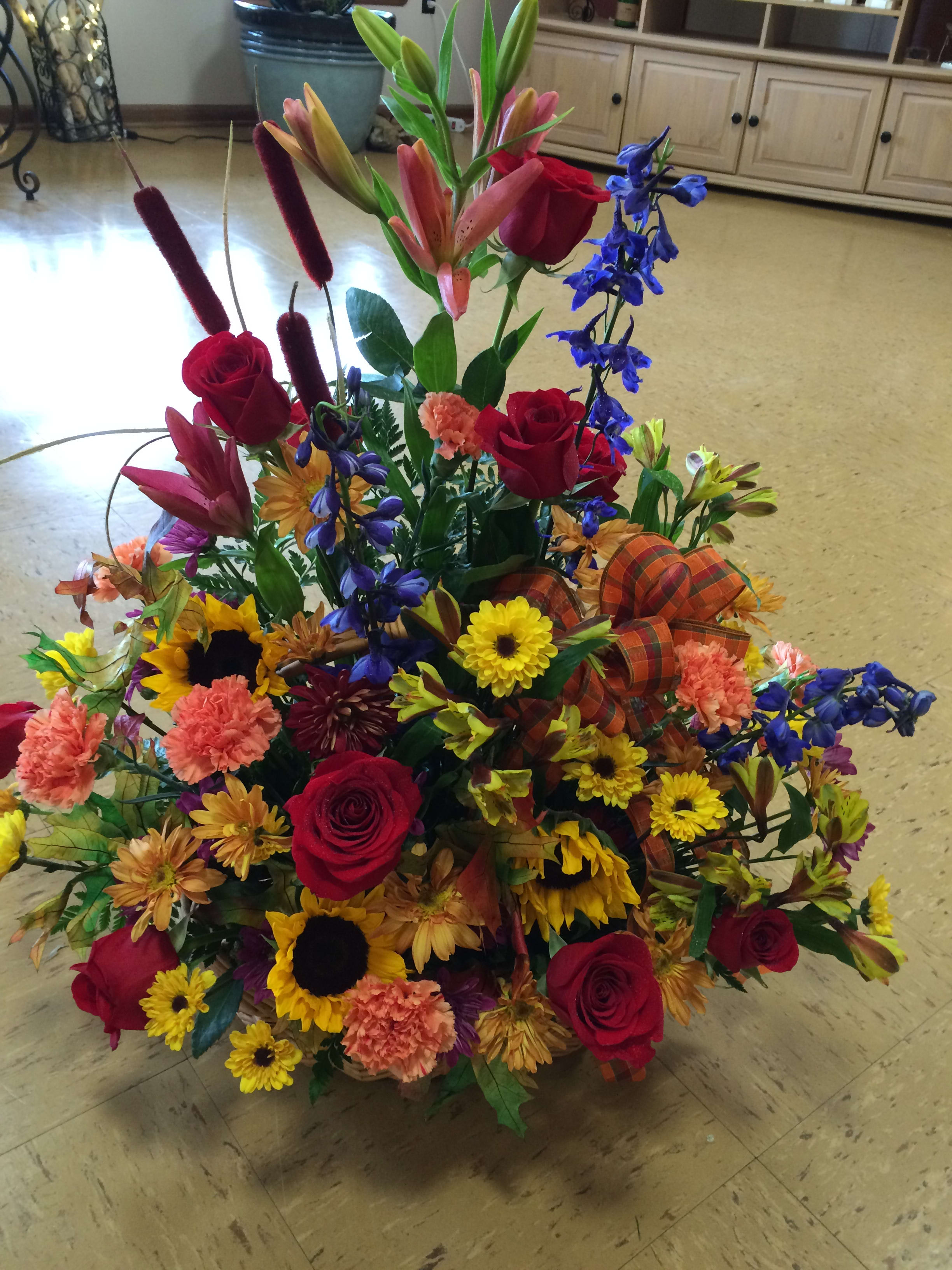 Autumn Garden - A mixture of fall colored flowers in reds, orange, peach, rust, yellow and dark blue. Delphinium, roses, and, sunflowers are the feature flowers.  Made in a wicker basket (if you prefer a vase, mention it in the special instructions box) This bouquet is great for almost any occasion, birthdays, thank you or as a condolence. (substitutions may apply)