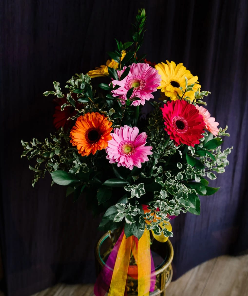 Gerbera Garden - Nestled within a sleek glass vase, a kaleidoscope of 12 gerbera daisies awaits. Picture vivid reds, sunny yellows, brilliant oranges, and cheerful pinks, each bloom exuding its own unique personality and radiance.