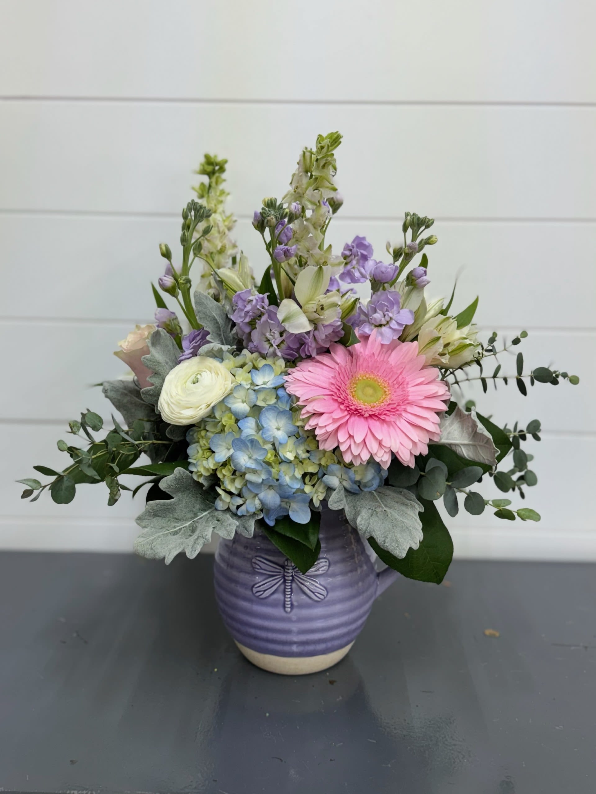 Pastel Dragonfly Pitcher Pastel Dragonfly Pitcher - With this beautiful whimsical vase is a stunning spring bouquet which includes, Ranunculus, Roses, Gerber Daisies, Alstromeria, Purple and White Stock, Dusty Miller, Hydrangeas, and other greenery assortment.