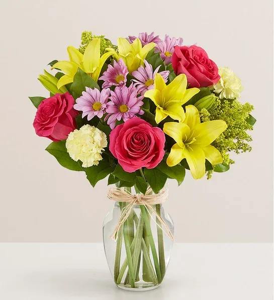 Fields of Europe for Mom Fields of Europe for Mom - A beautiful bouquet of vibrant flowers arranged in a clear glass vase . The arrangement features several bright pink roses , cheerful yellow lilies , delicate light purple daisies , and fluffy white carnations . Lush green leaves provide a lovely backdrop. The vase is tied with a simple raffia bow . The overall impression is one of freshness and joy .