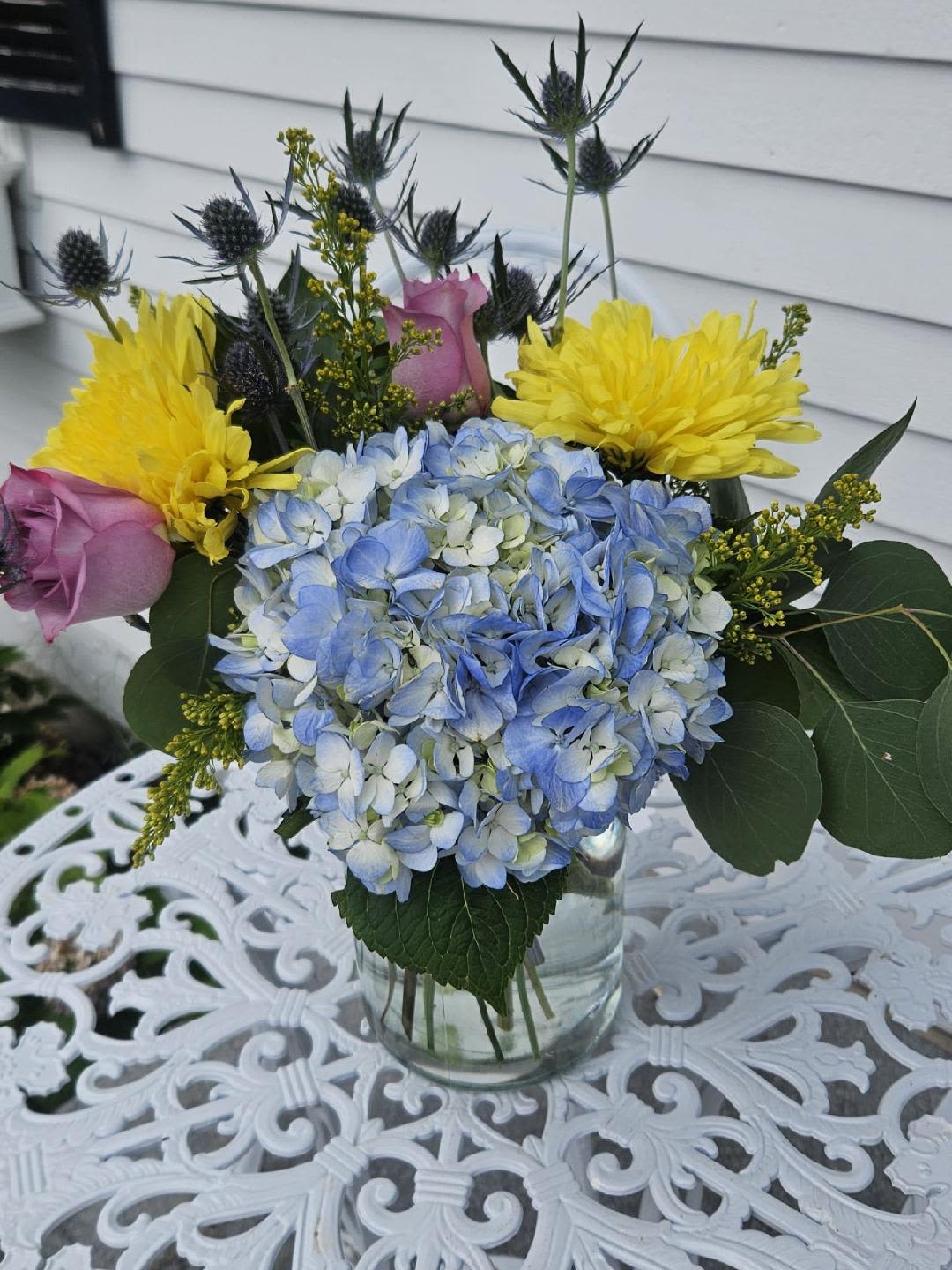 Deal of the Day Flower Shop - Blue hydrangea, lavender roses, yellow cushion mums blue thistle, solidago and dollar eucalyptus arranged in a  glass vase
