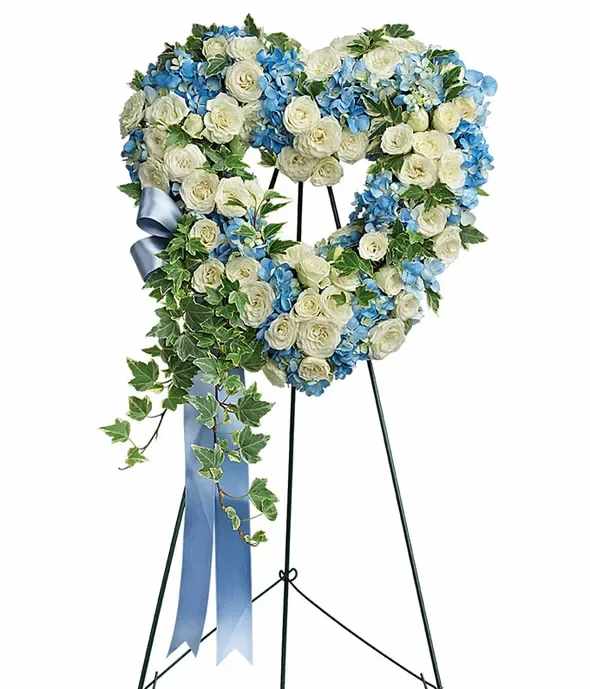 Heartfelt Serenity Funeral Spray - A heart-shaped funeral spray elegantly designed with a blend of soft white roses and delicate blue hydrangeas. Accented with lush green ivy, the arrangement is complemented by a cascading blue ribbon, creating a serene and comforting tribute. Displayed on a sturdy metal easel, this spray offers a heartfelt expression of love and sympathy.