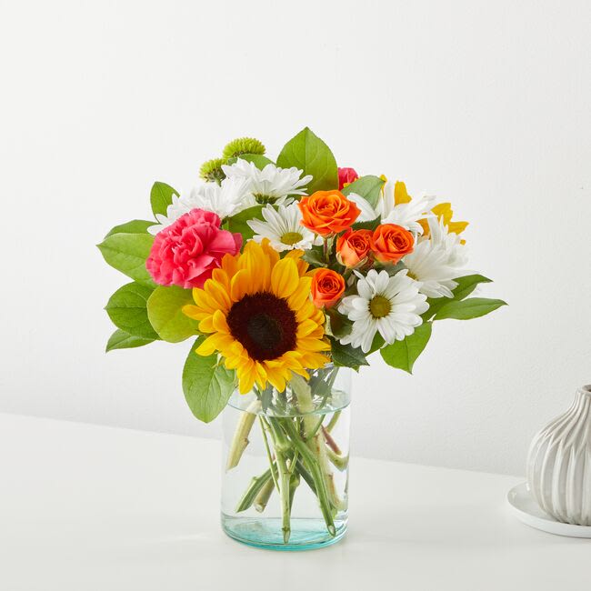 Sun-Drenched Blooms Bouquet - Welcome warmer weather with the Sun-Drenched Blooms Bouquet! Sunflowers, hot pink carnations, white daisies, orange spray roses, and green button pompons celebrate the joy of the season. Vase style may vary!