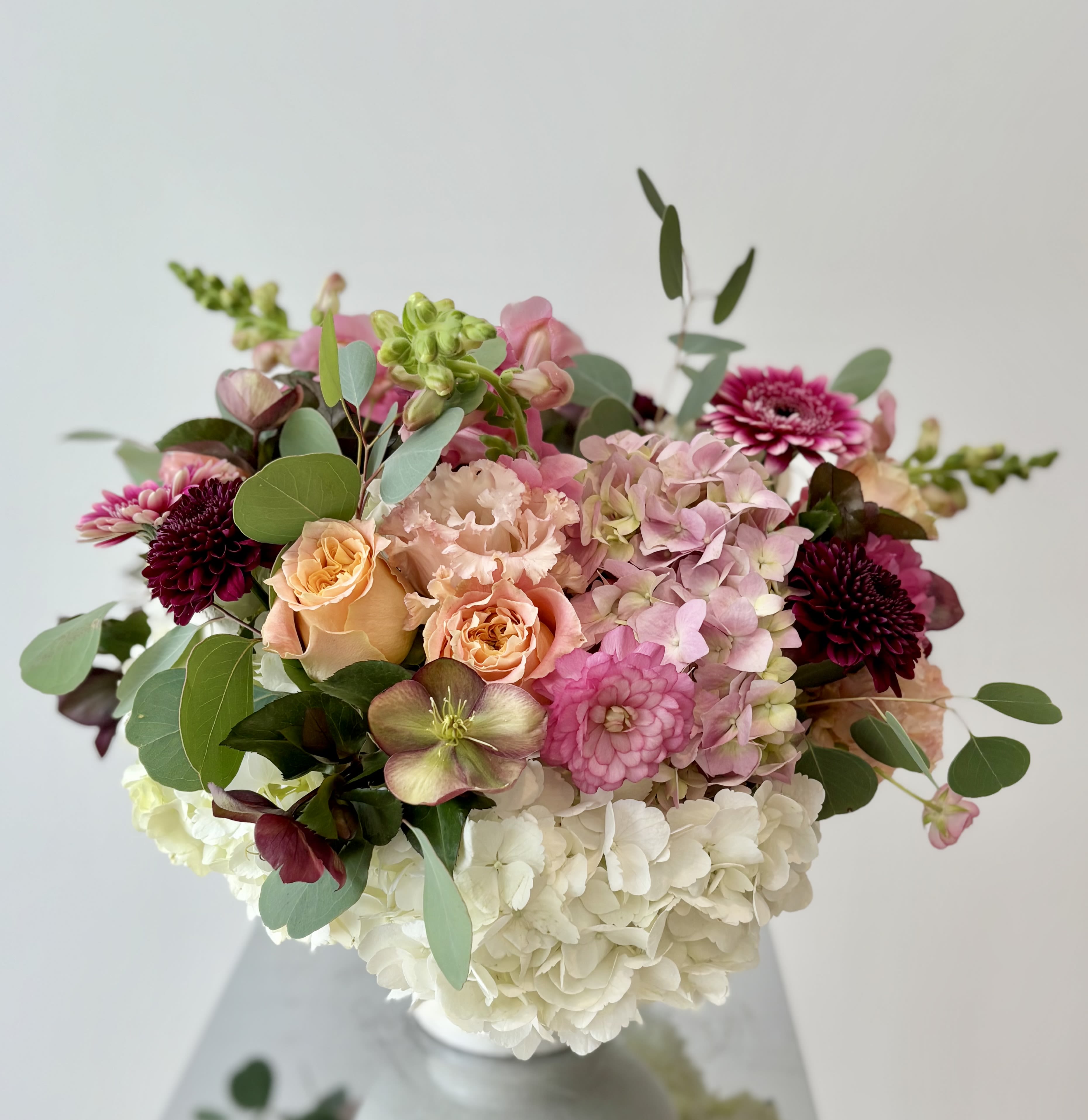 All Yours - A lovely flower centerpiece in a compote vase with premium flowers the pink and red/burgundy color shades nestled among white hydrangeas, with a pop of peachy blooms and green foliage. 