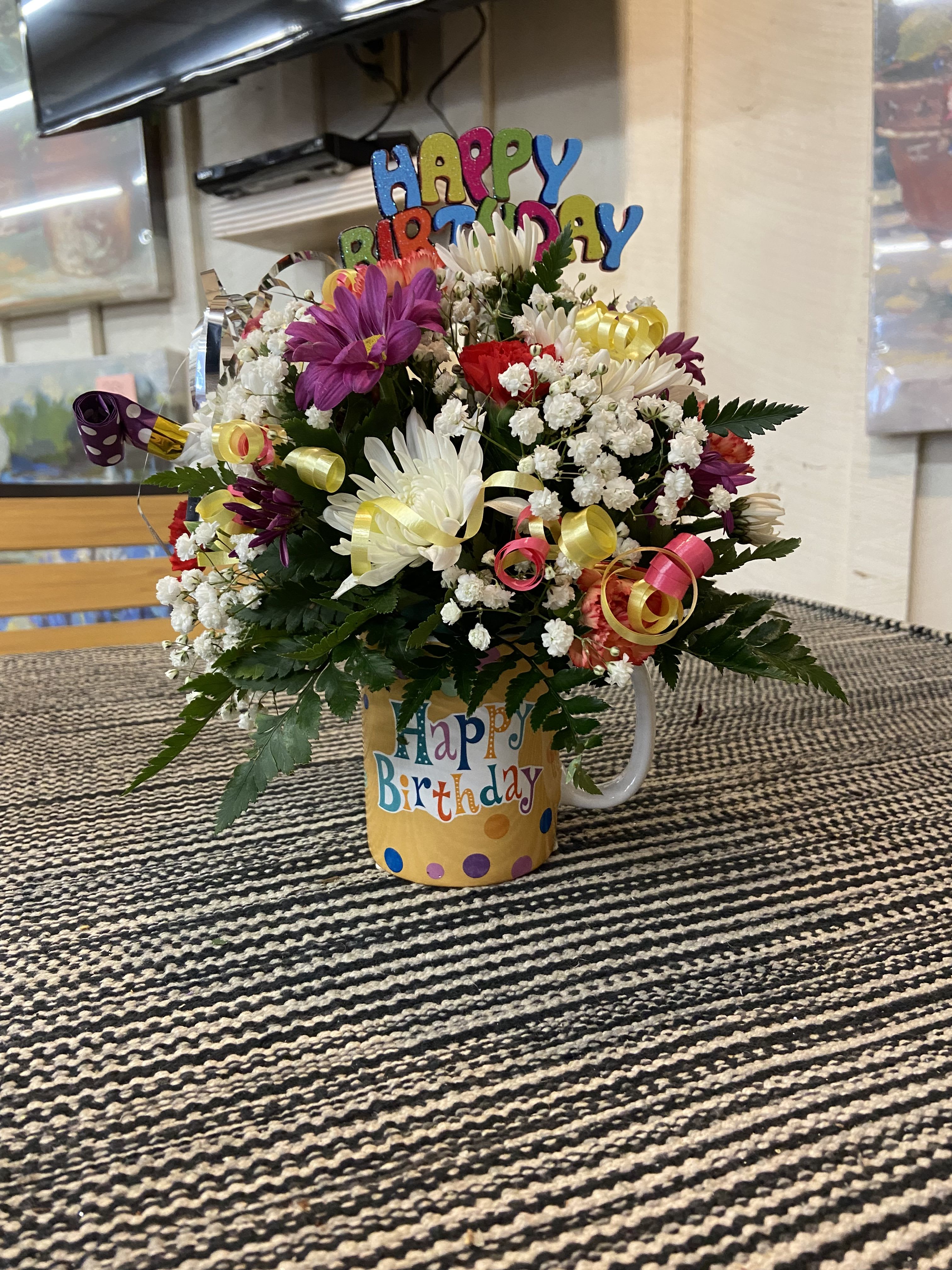Birthday Mug Birthday Mug - Time to celebrate with this super fun arrangement designed in a "Happy Birthday" mug filled with flowers in the colors of red, yellow, orange, and purple. Topped with ribbon, happy birthday picks, and party horns.