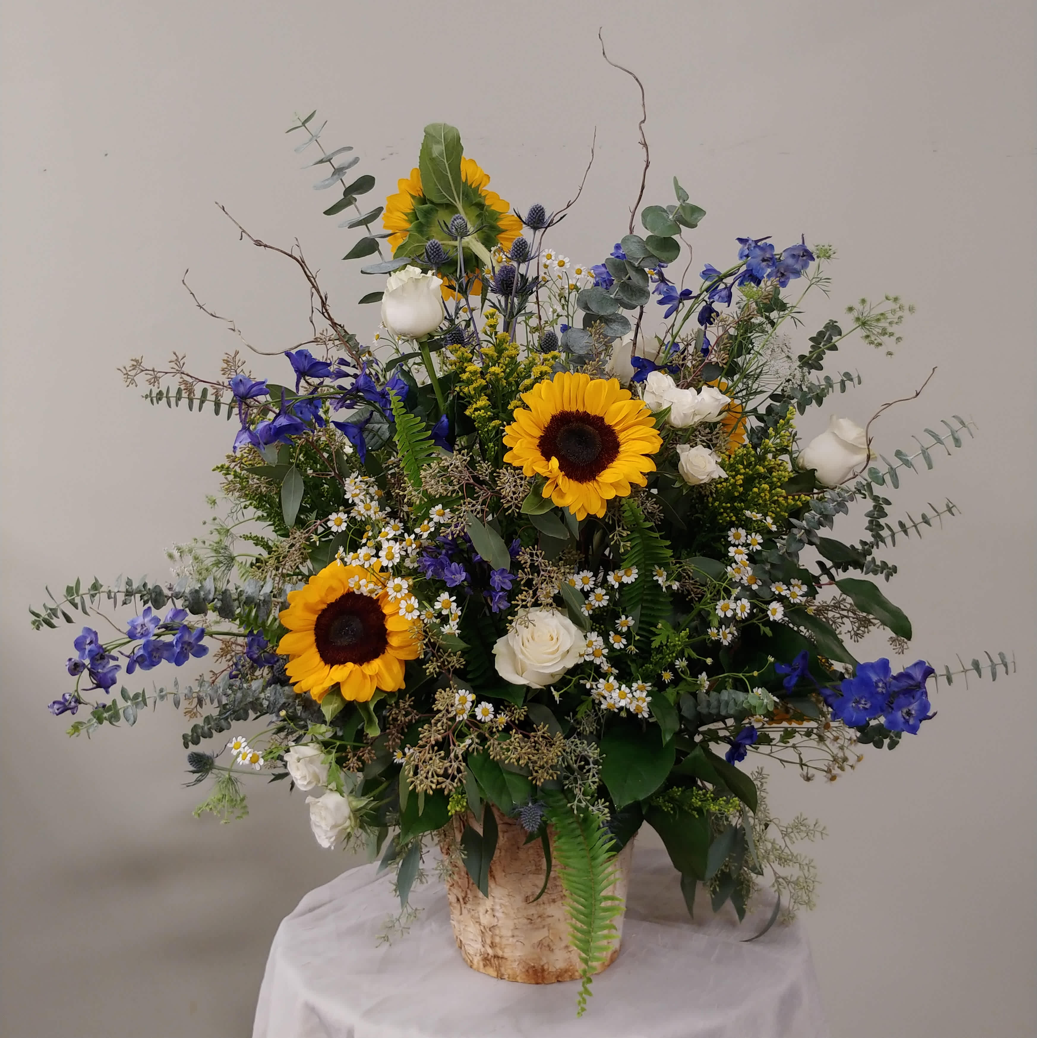 Sunflower and Blue Delphinium Floor Basket Sunflower and Blue Delphinium Floor Basket - A large birch container featuring Sunflowers, white Roses, blue Delphinium, seasonal complementary florals and greens. Although you can place memorial orders online, we recommend you call the store to ensure we achieve your vision.