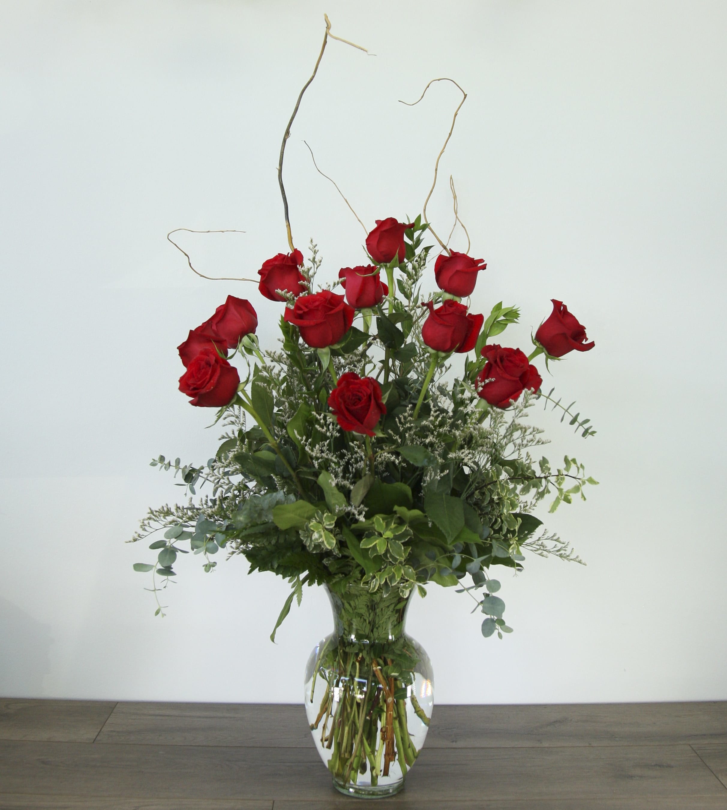 Classic Red Roses- With A Twist! (EXP 22) - The classic red rose arrangement for romantic occasions, with a twist! In this rendition of red roses, we've added curly willow and silver dollar eucalyptus is also included for more added volume, length, and texture. The arrangement is completed with an assortment of fresh greenery for added dimension and color. Placed inside a curved bottom-heavy glass vase. 