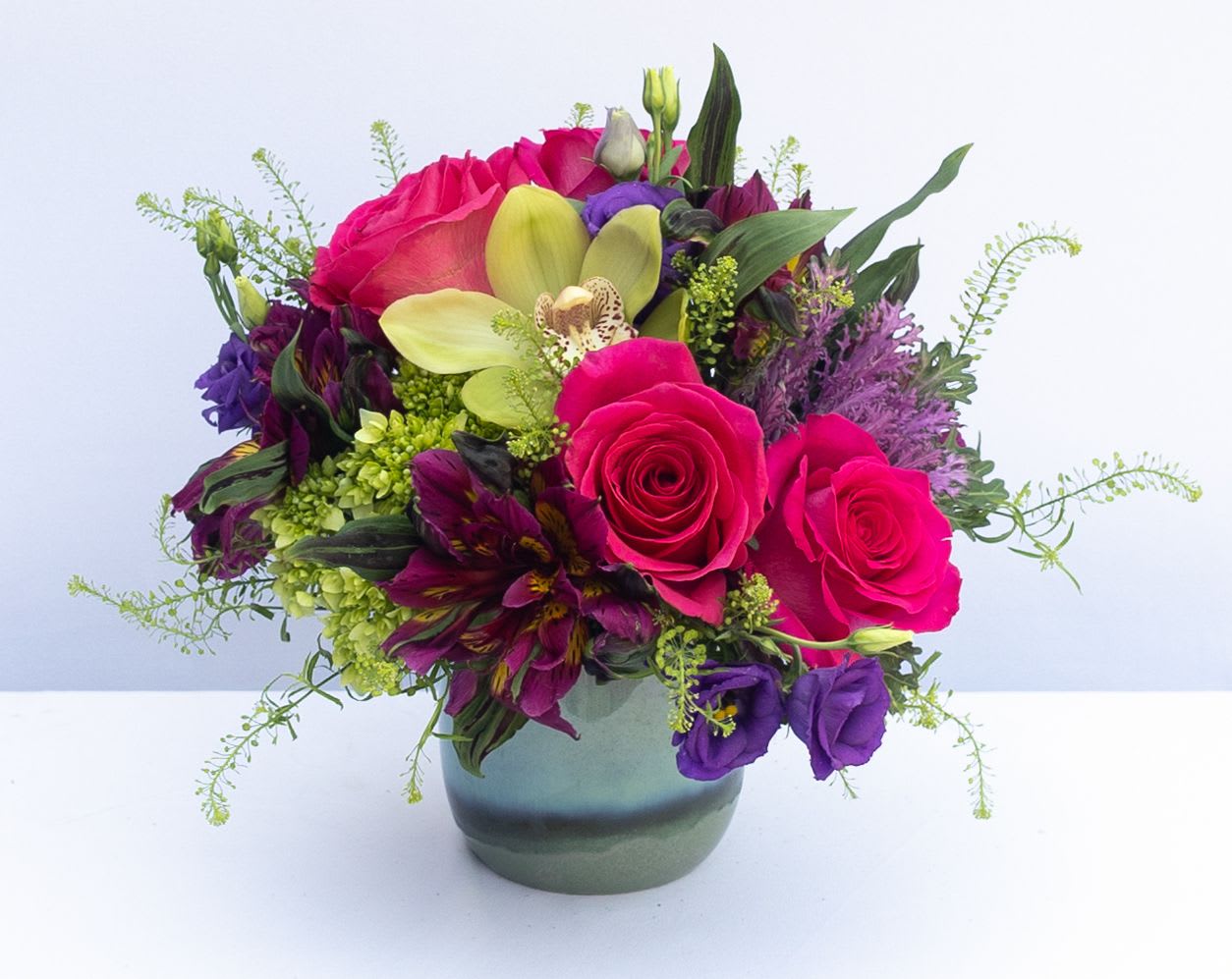 Petite Jewel Tones Petite Jewel Tones - Small arrangement in a small blue ceramic vase. Flowers may include hot pink roses, vibrant purple alstroemeria, Purple Lisianthus, and cymbidium orchid.