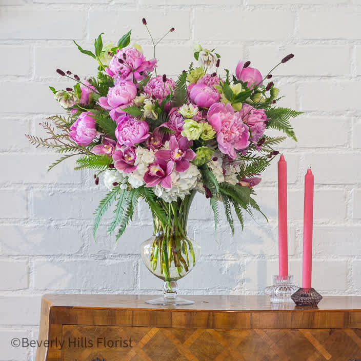 Petals of Elegance  - Behold the exquisite beauty of our Floral Fusion arrangement, delicately showcased within a glass case. Lush peonies and cymbidium orchids intertwine with white hydrangeas in a graceful dance of elegance and charm. Housed in a short yet tall glass vase, this ensemble exudes a timeless allure, captivating the senses with its harmonious blend of colors and textures. Perfect for adding a touch of sophistication to any space, let this arrangement elevate your surroundings with its enchanting beauty and grace.