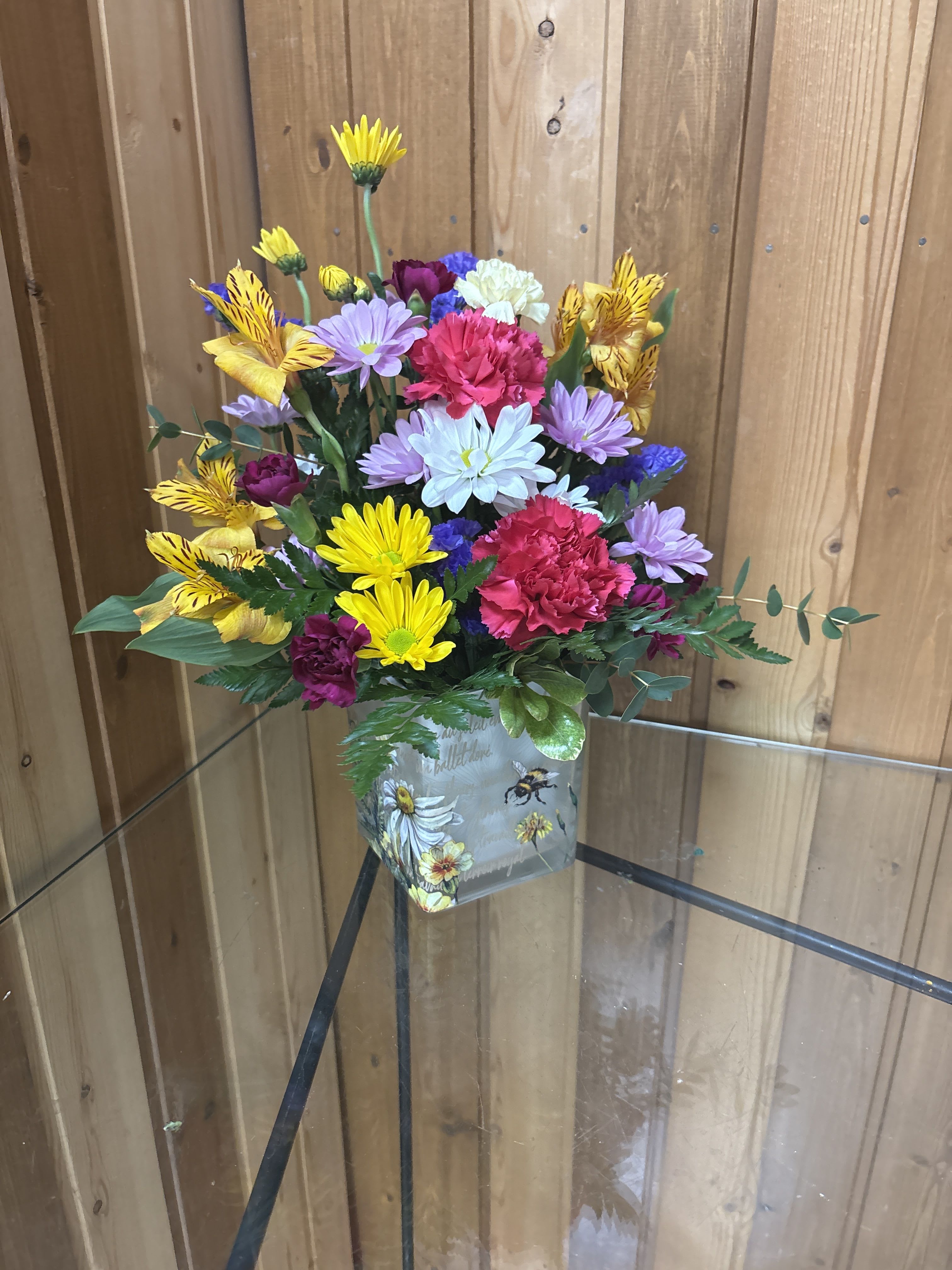 Floral Buzz  - Like a bee dancing through a hidden garden, this enchanting bouquet of orange, lavender, and yellow blooms in a vintage-inspired glass cube makes for a truly magical gift. This delightful arrangement features vibrant orange alstroemeria, lavender carnations, miniature pink carnations, yellow daisy spray chrysanthemums, and pink sinuata statice, all beautifully accented with leatherleaf fern and pitta negra. It is presented in Teleflora's Floral Buzz Cube, making it a charming addition to any tablescape. Orientation: All-Around  SUBSTITUTION POLICY – Always deliver the freshest flowers! Please note the bouquet pictured reflects our original design.  If the exact flowers or container in this arrangement are not available, our local florists will create a beautiful bouquet with the freshest available flowers.