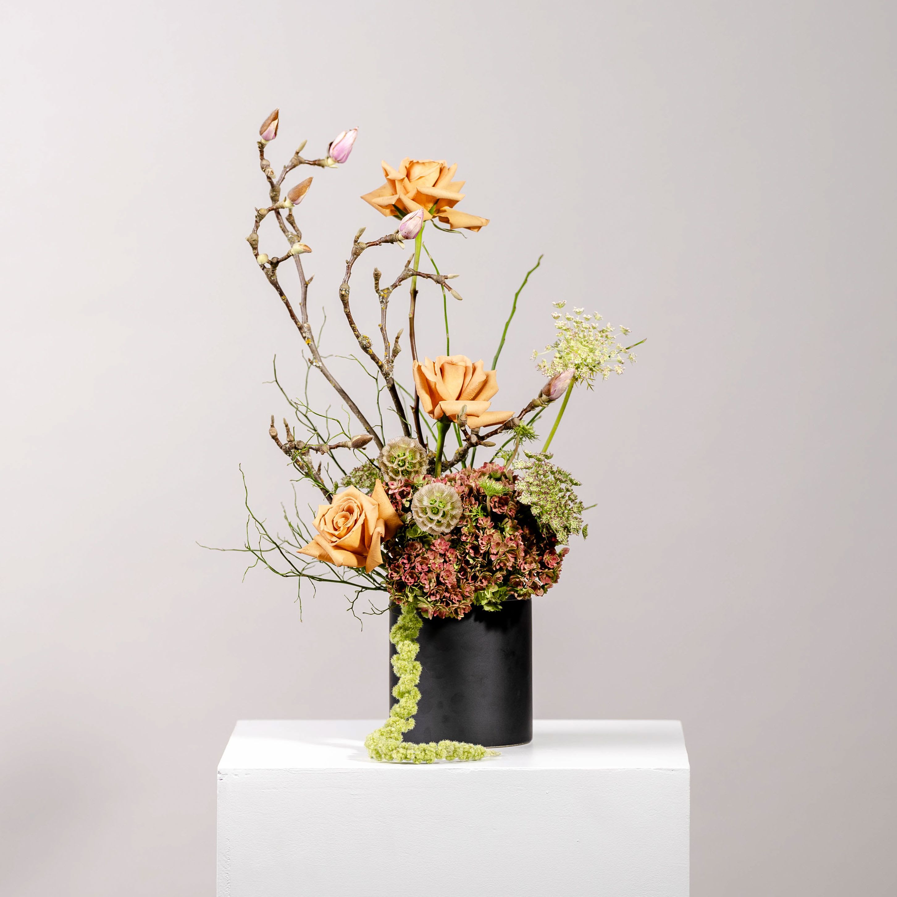 Seasons of Love - A striking floral arrangement in a sleek black vase. Warm tan roses take center stage, complemented by the earthy tones of hydrangeas and branches that add height and structure. Delicate pink buds introduce a hint of romance, while trailing green vines bring a touch of life and movement.   It’s a sophisticated blend of textures and colors—ideal for adding elegance to any space or making a bold statement in your home.   *Picture shown is of DELUXE arrangement
